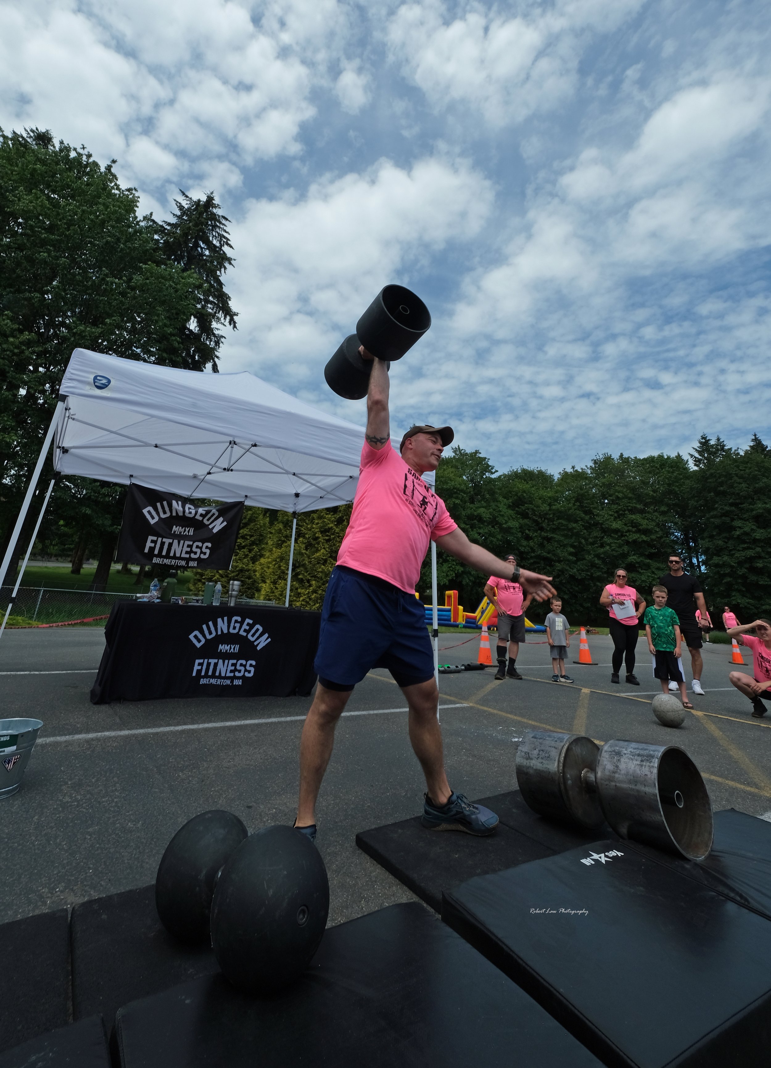 Man lifting heavy dumbbell overhead at outdoor fitness event with spectators and tent in background.