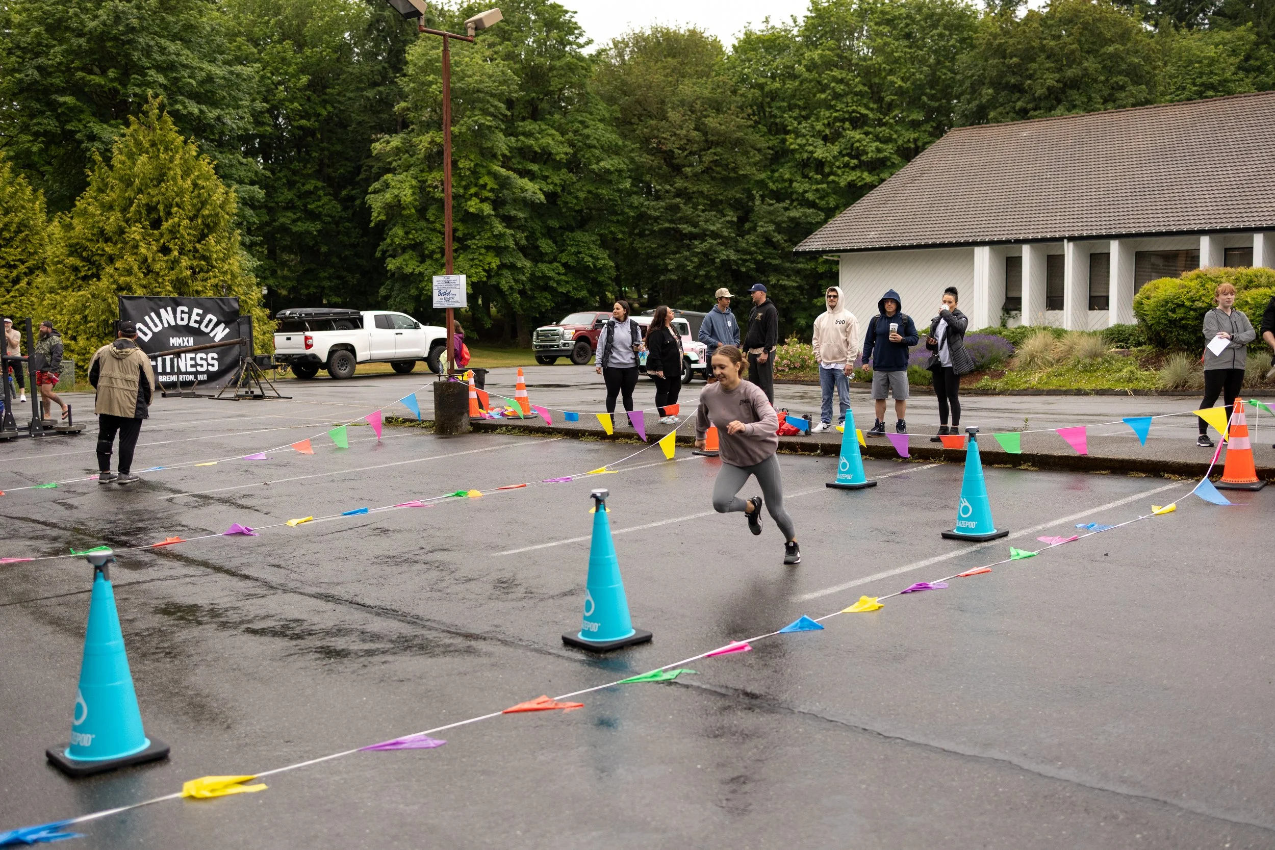 A girl running in a race in a parking lot, which is decorated with colorful cones and banners. Several spectators are watching and taking photos—some bundled up due to rainy weather. Bandstand with 