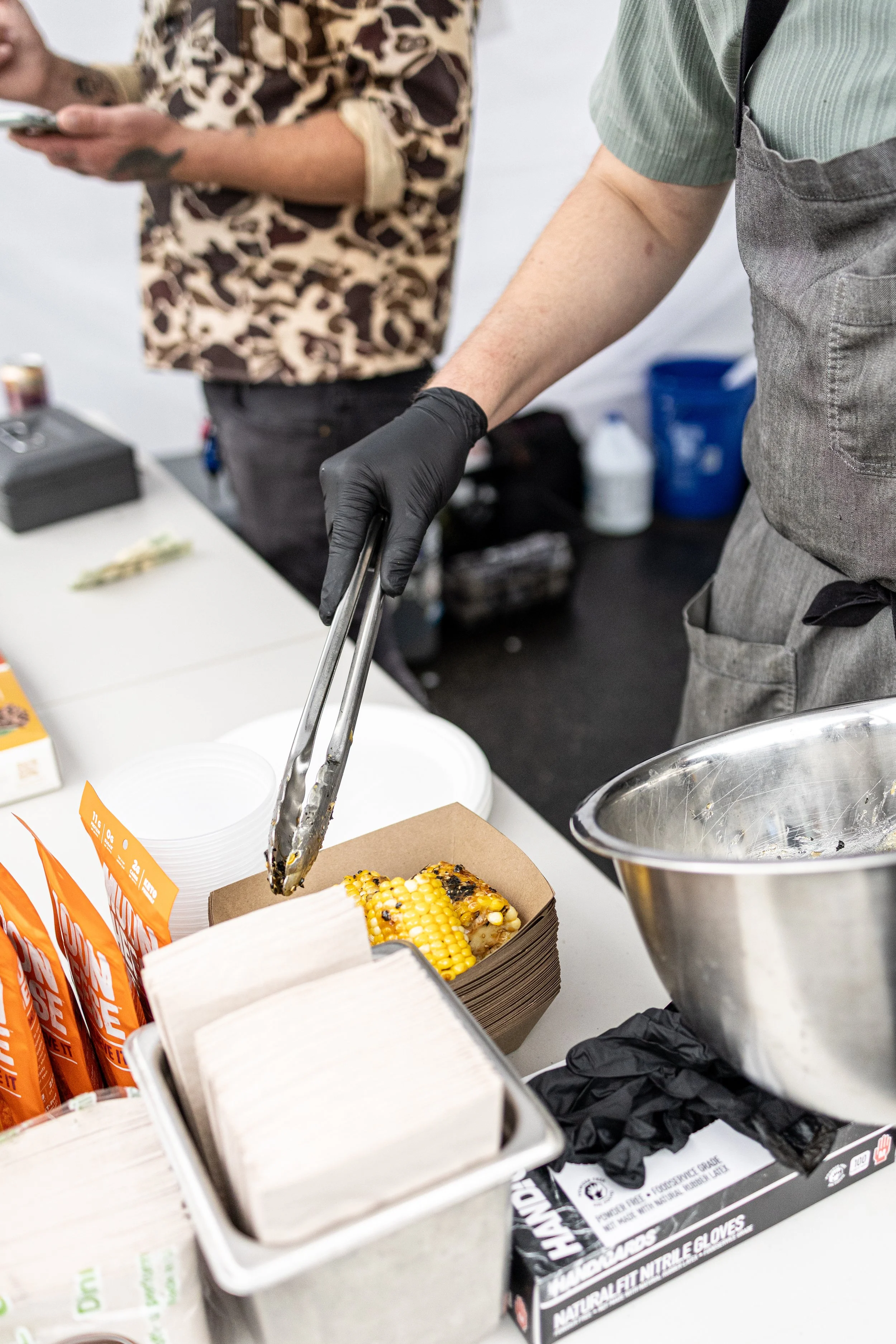 Person wearing black gloves is serving grilled corn on the cob with tongs at a food station, with another person in a leopard print shirt in the background.