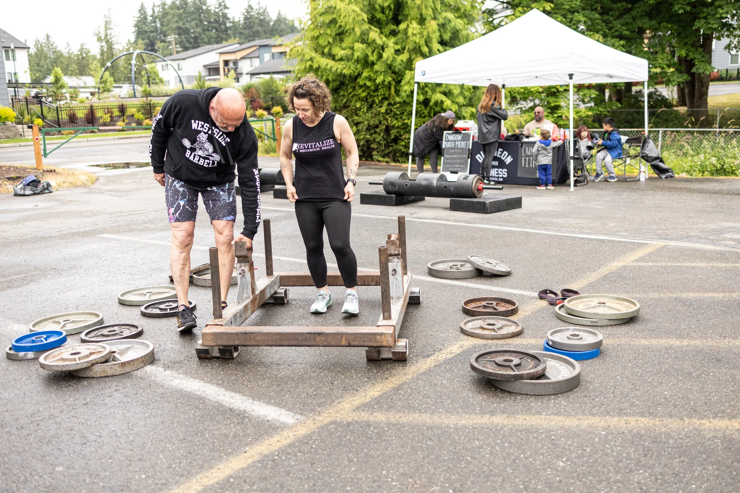 People lifting weights outdoors in a parking lot, with a tent and tables in the background, and various weight plates scattered on the ground.