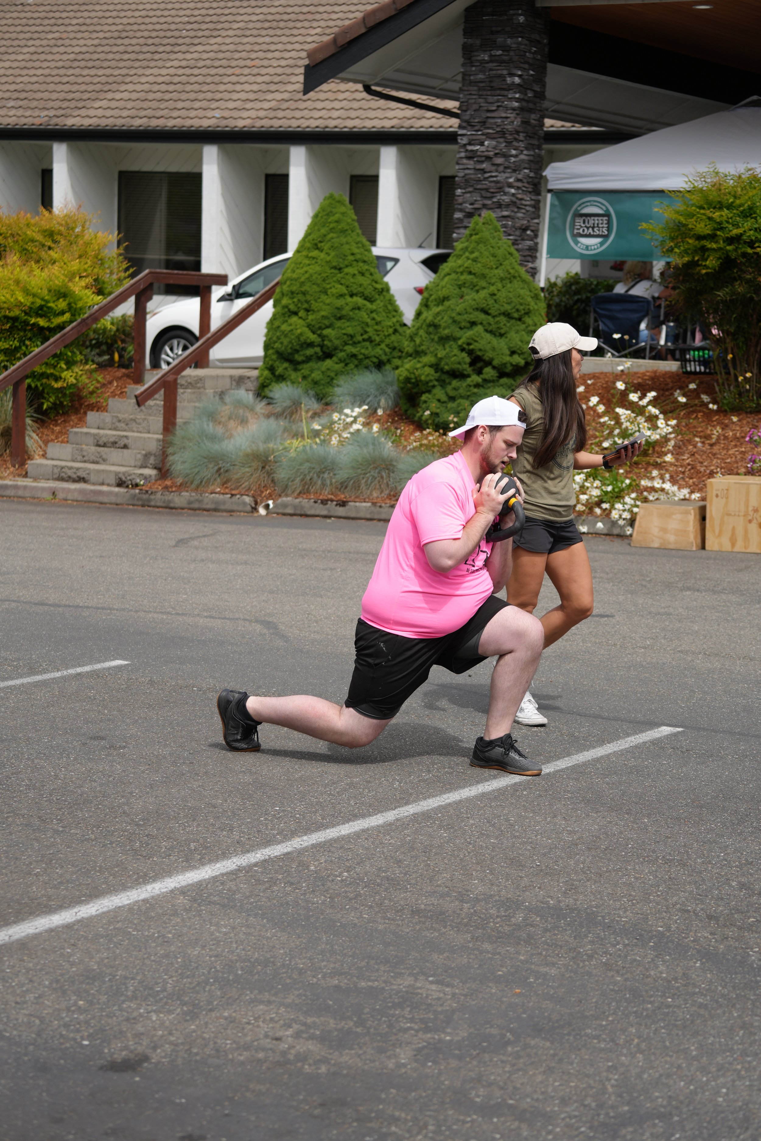 A man in a pink shirt and black shorts doing lunges with a kettlebell in a parking lot next to a woman with a phone.