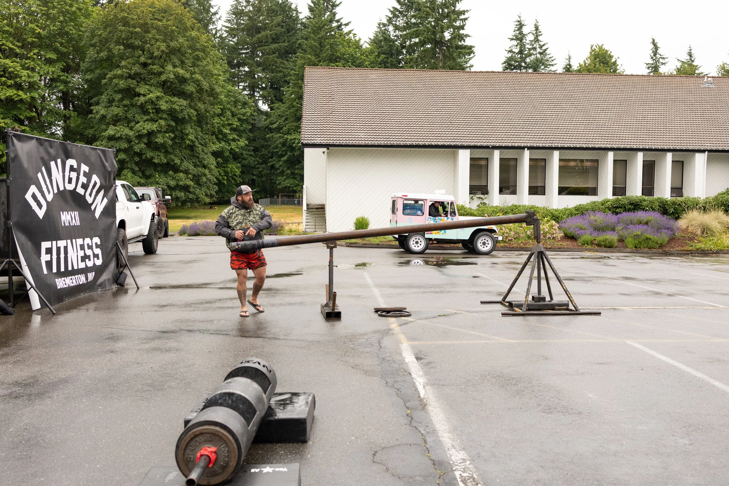 Man preparing for a strongman lift outdoors in a parking lot, with a large black banner reading 'Dungeon Fitness Bremerton, WA' on the left and weightlifting equipment nearby, during a rainy day.