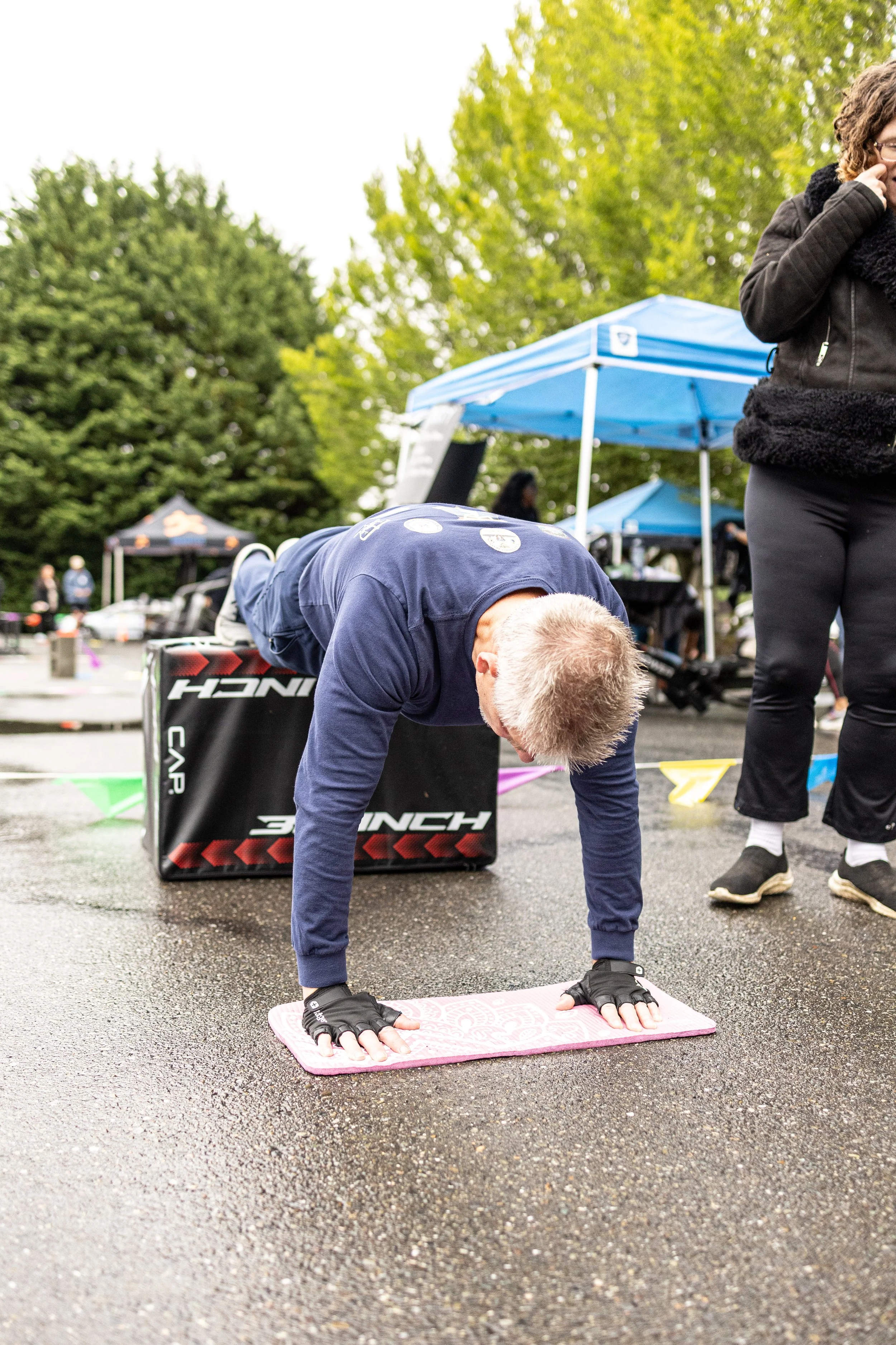 Man performing a push-up on a pink yoga mat outdoors during a fitness event with tents and trees in the background.