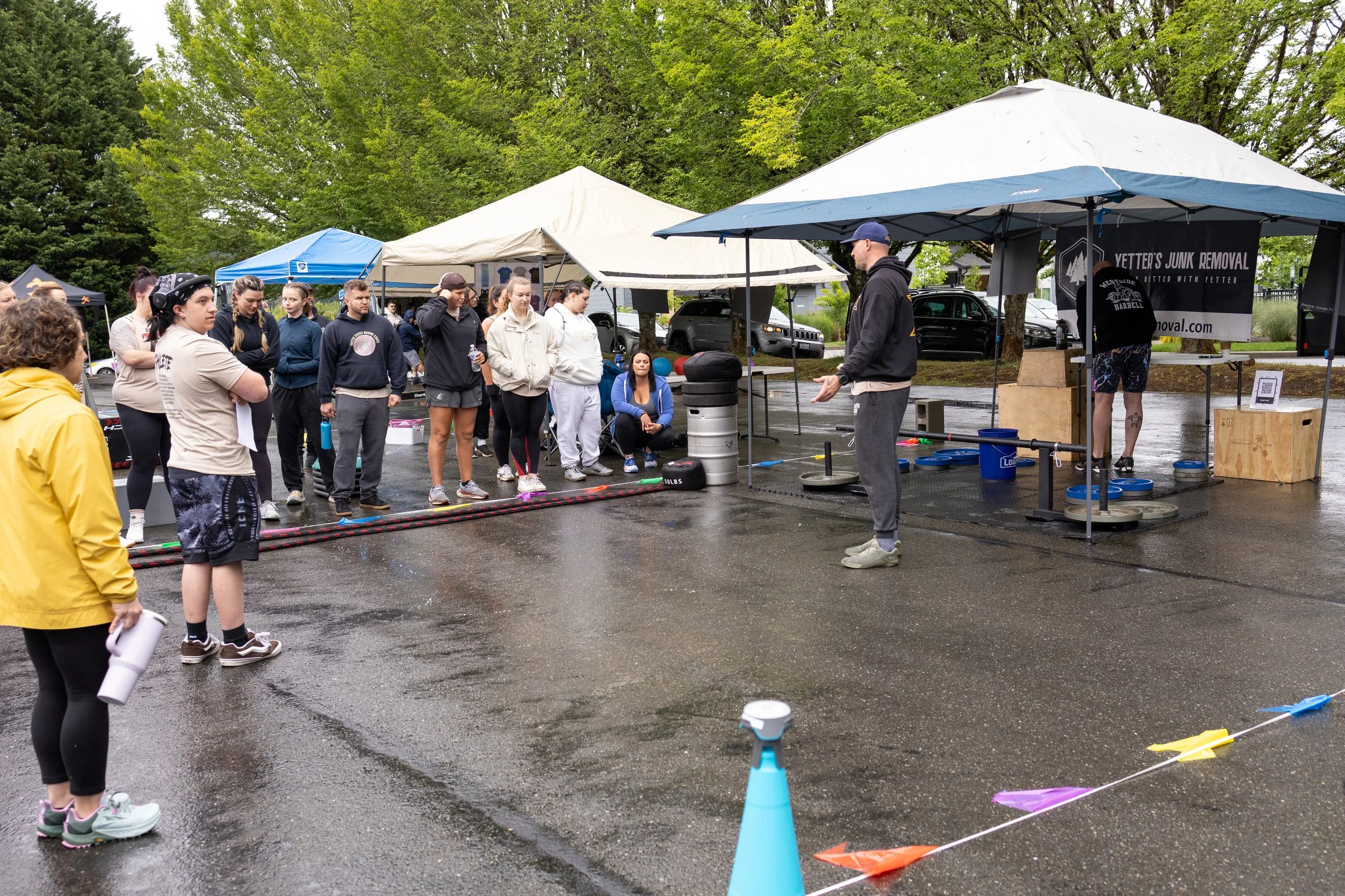 People gathered outdoors near tents for a fitness class or demonstration, with an instructor explaining while others watch and listen, on a wet day with trees in the background.