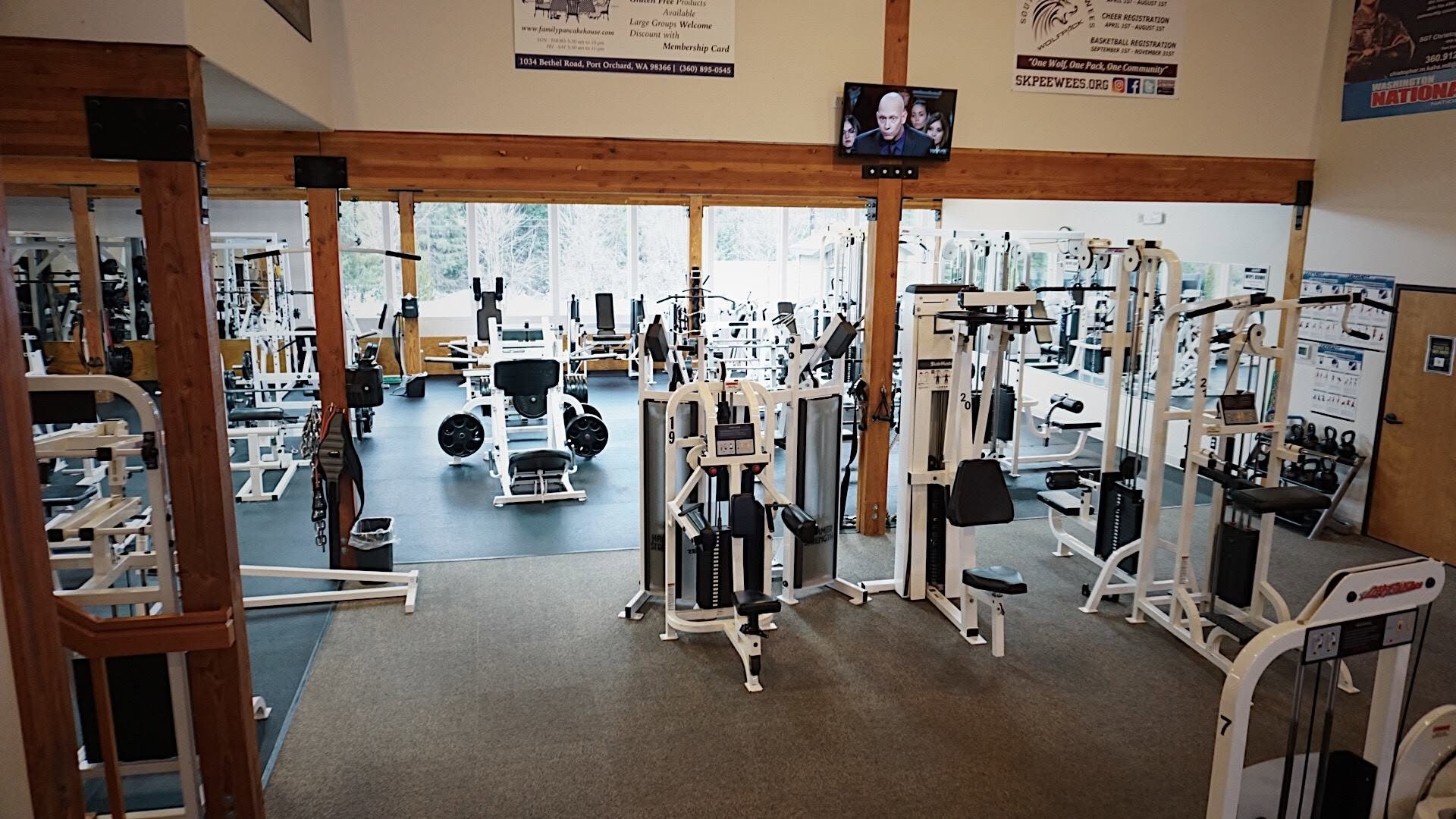 Interior of a gym with various white workout equipment and machines, large windows on the back wall, and sports posters on the walls.