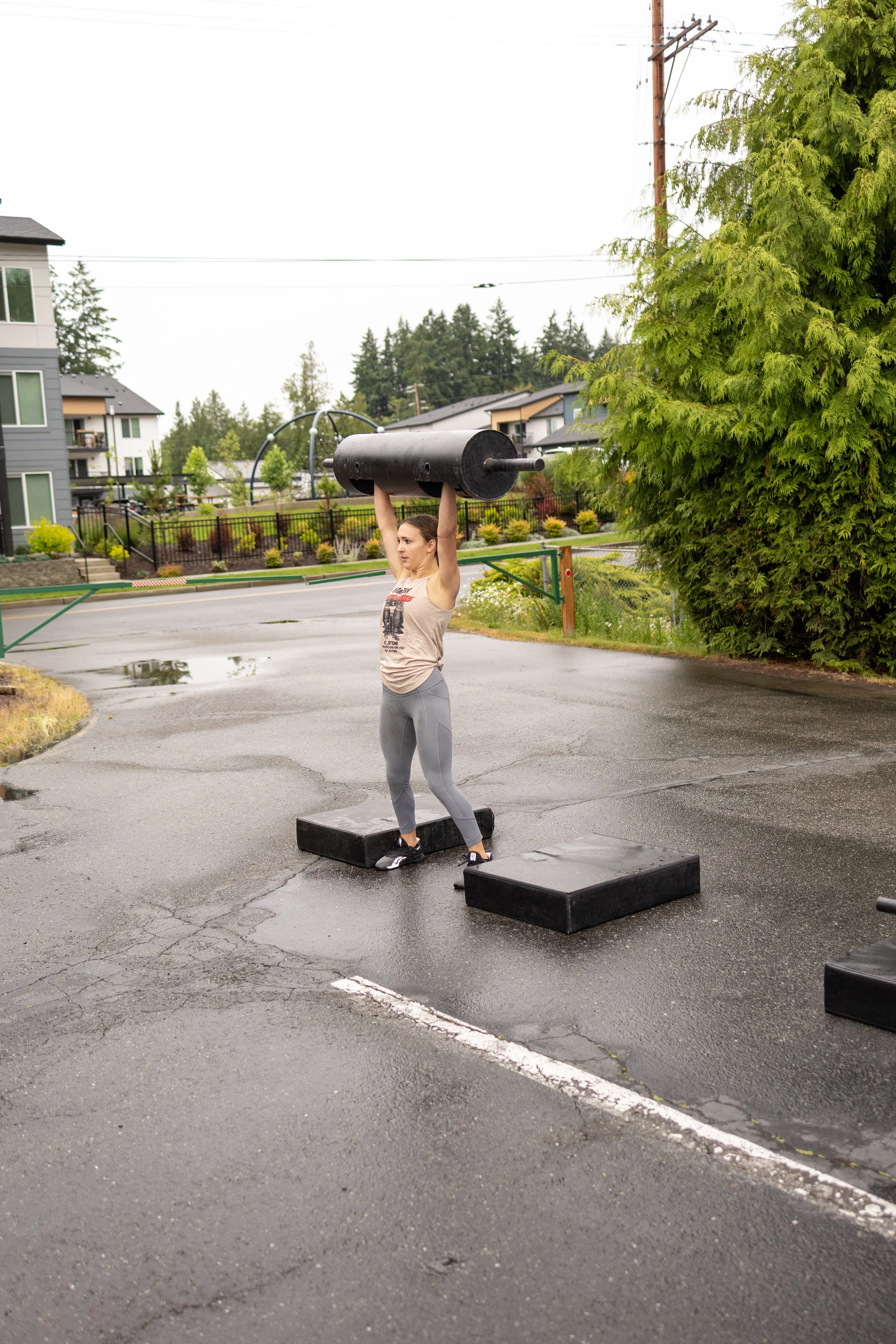 A woman lifting a large black weight over her head outdoors in a parking lot on a cloudy, wet day.