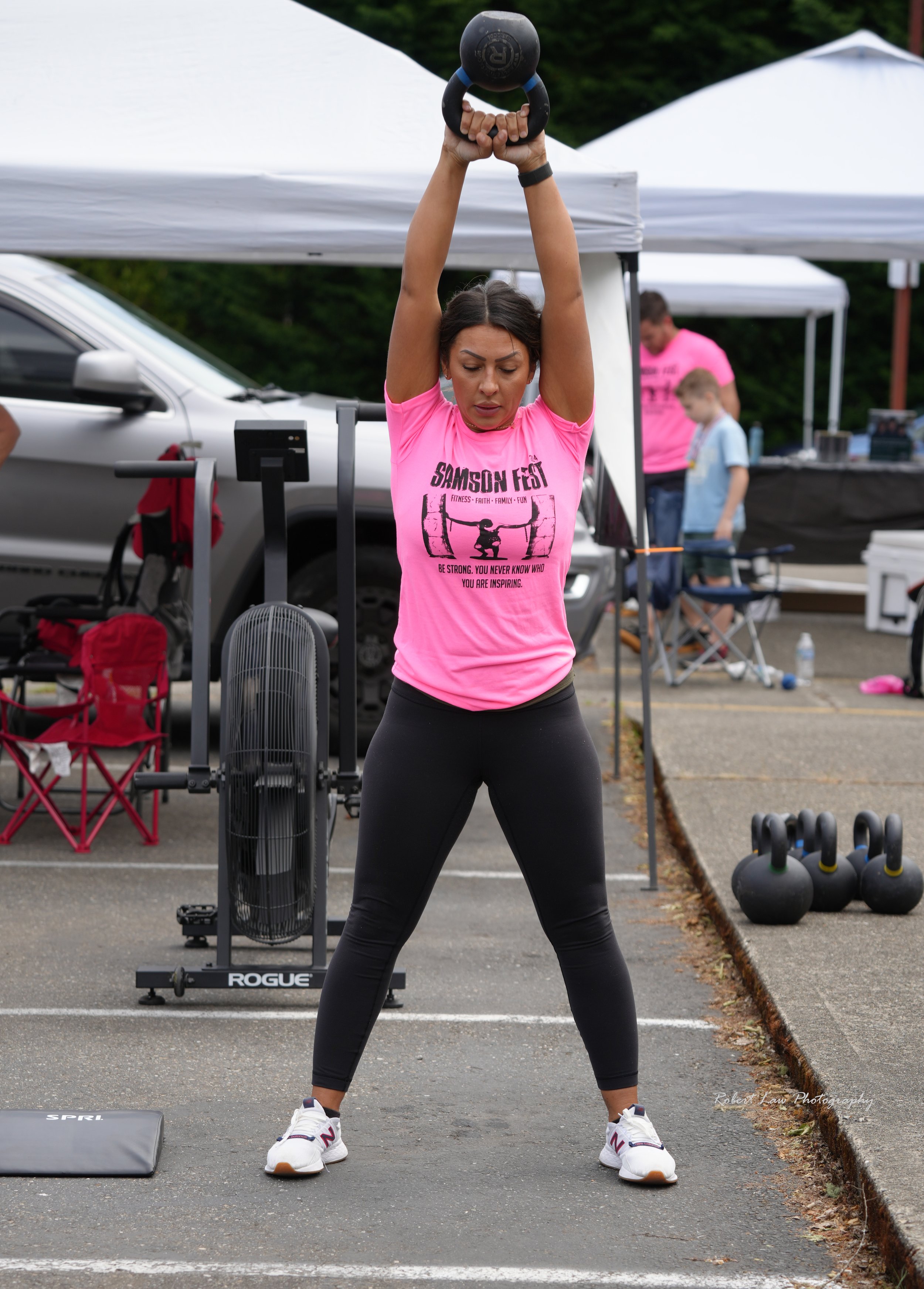 A woman in a pink shirt performing a kettlebell swing outdoors at a fitness event. Workout equipment and tents are visible in the background.