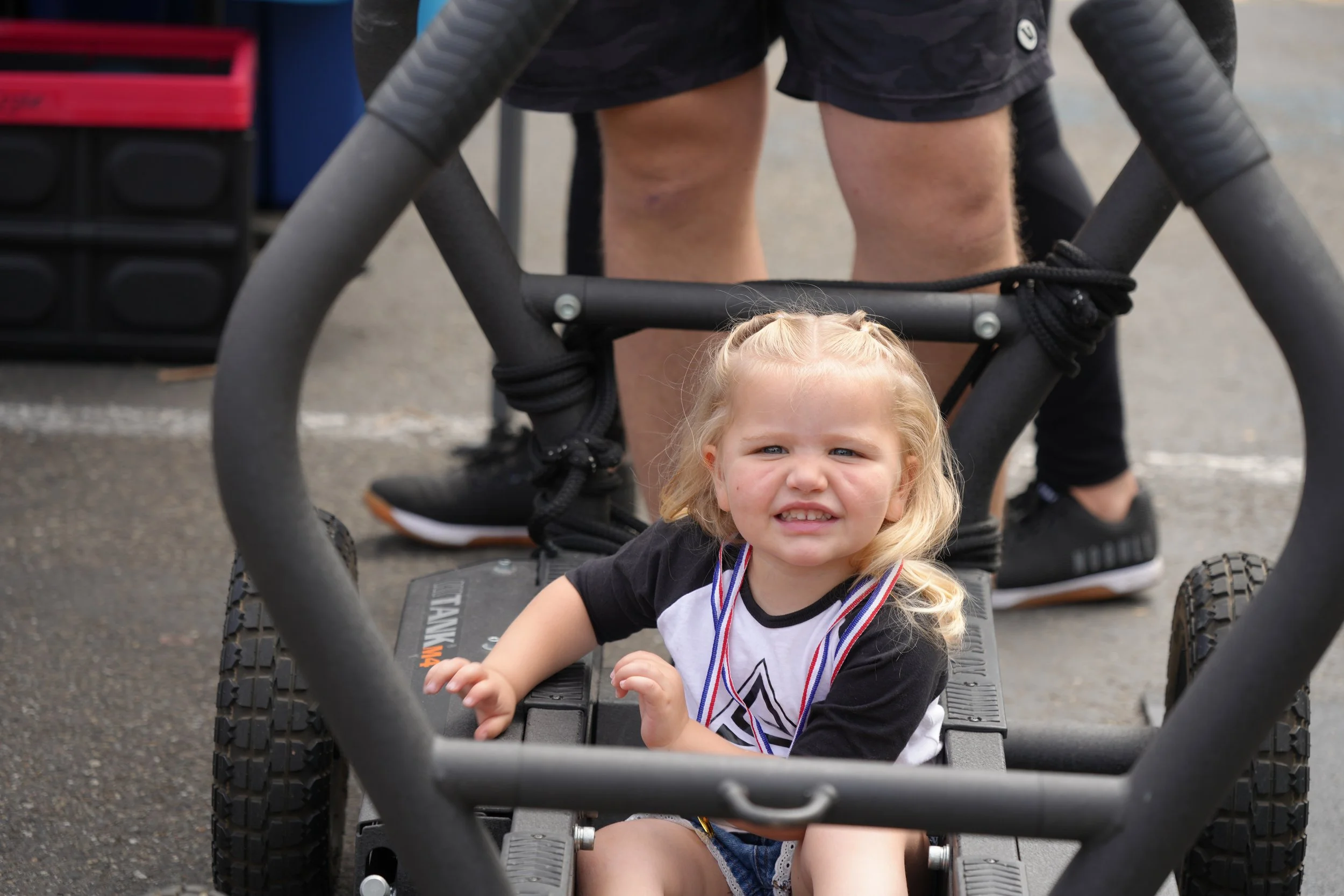 Young child sitting in a pushcart, smiling outdoors; adult legs visible in the background.