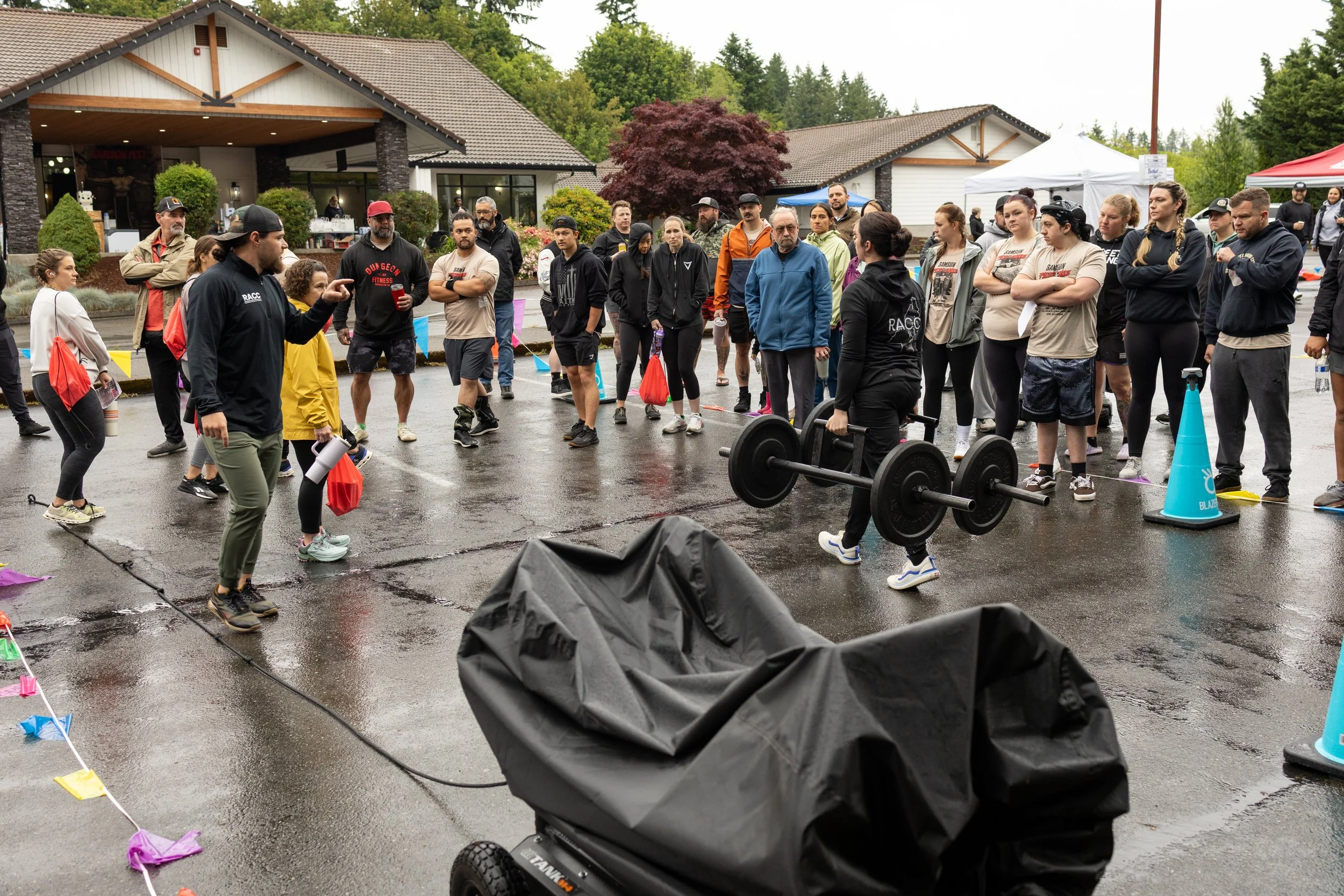 A group of people gathered outdoors on a wet, overcast day, participating in or watching a fitness event. A woman is carrying a barbell, and a man is speaking to the group. The setting appears to be outside a building with tents and trees in the back