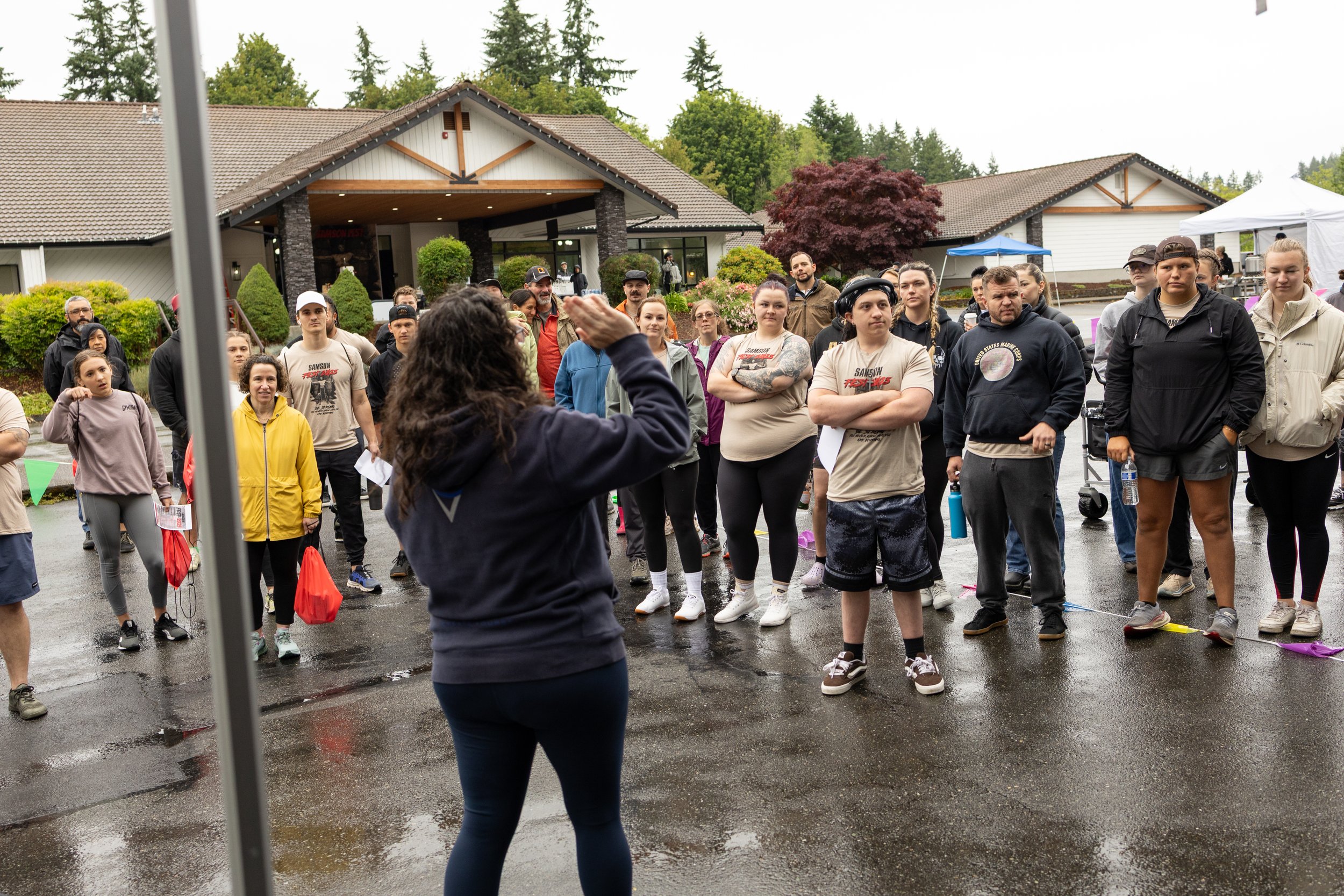 A woman in a dark jacket appears to be speaking or instructing a group of people gathered outside, with a building and trees in the background.