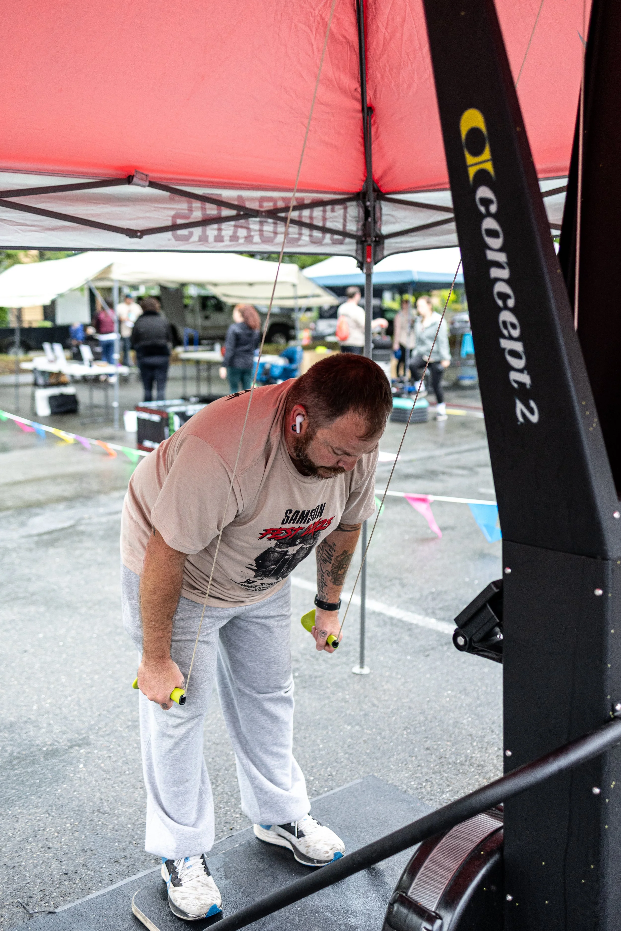 A man participating in a fitness event, holding a jump rope and using a rowing machine outdoors under a red canopy.