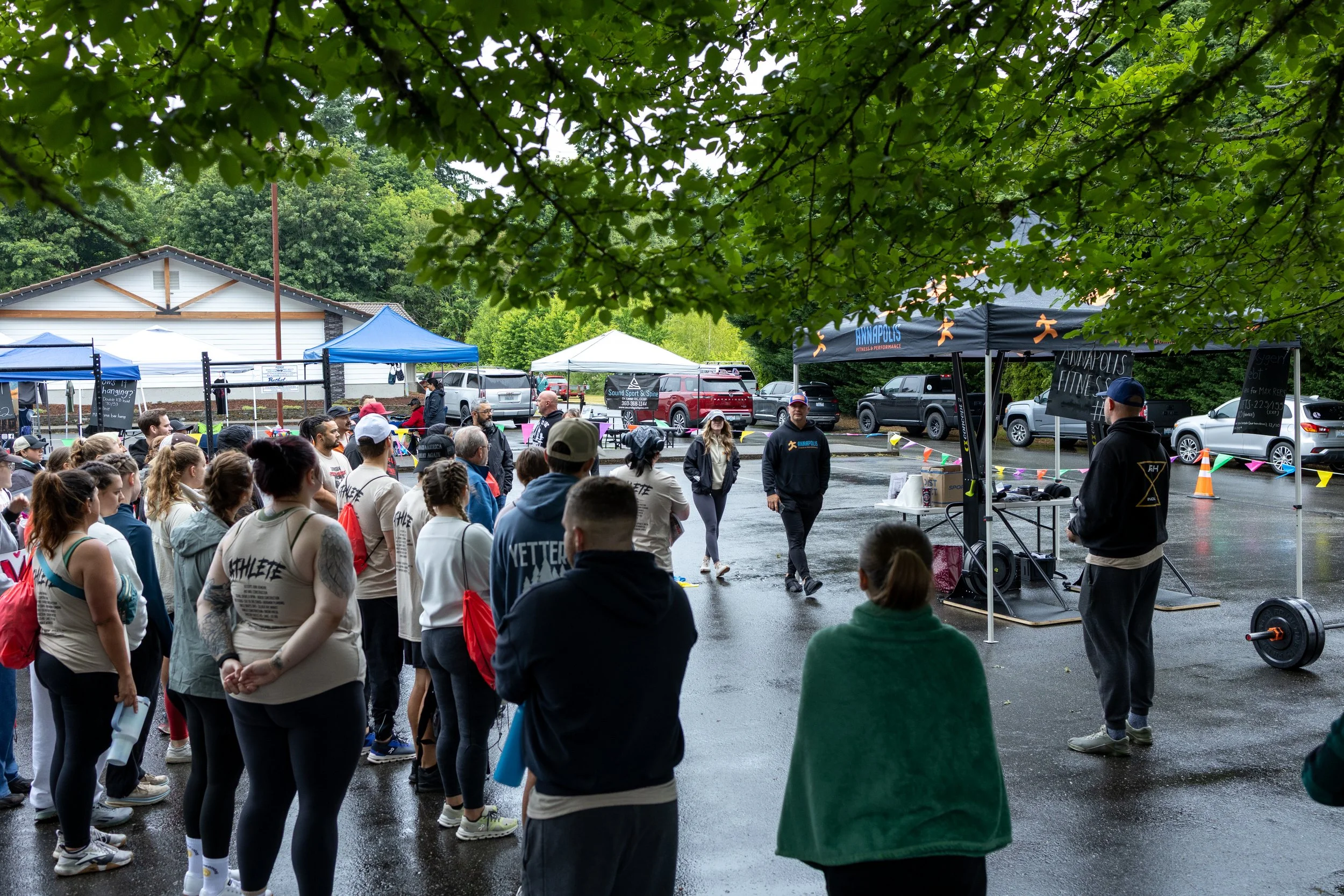 A group of people gathered outdoors in a parking lot for a fitness event, with tents, cars, and trees in the background, and a person speaking to the crowd under a canopy.