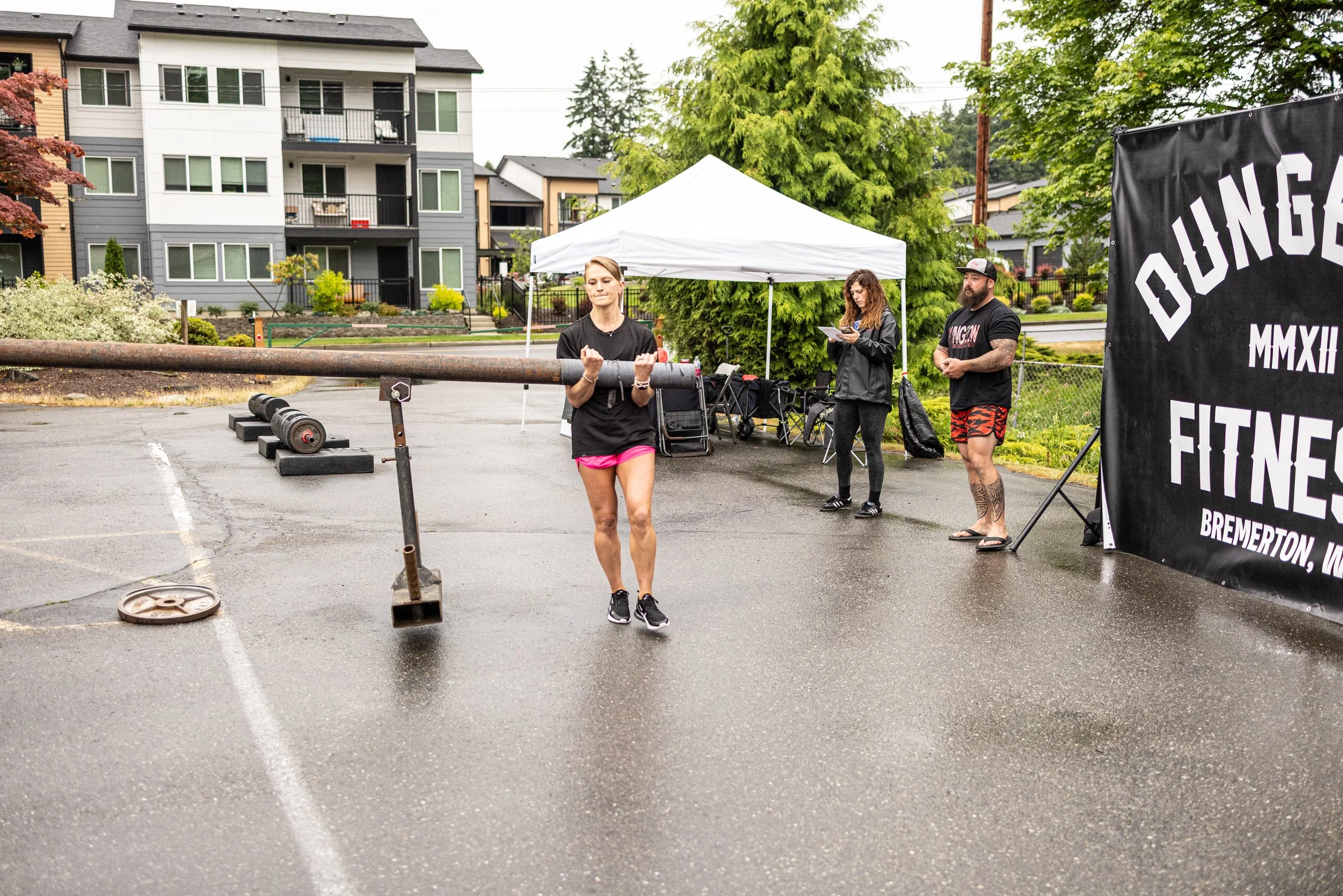 A woman lifting a large metal bar outside during a workout event, with two people standing nearby and a banner that reads 'Dunga Fitness' and 'Bremerton, WA' under a white canopy, in a parking lot with residential buildings in the background.