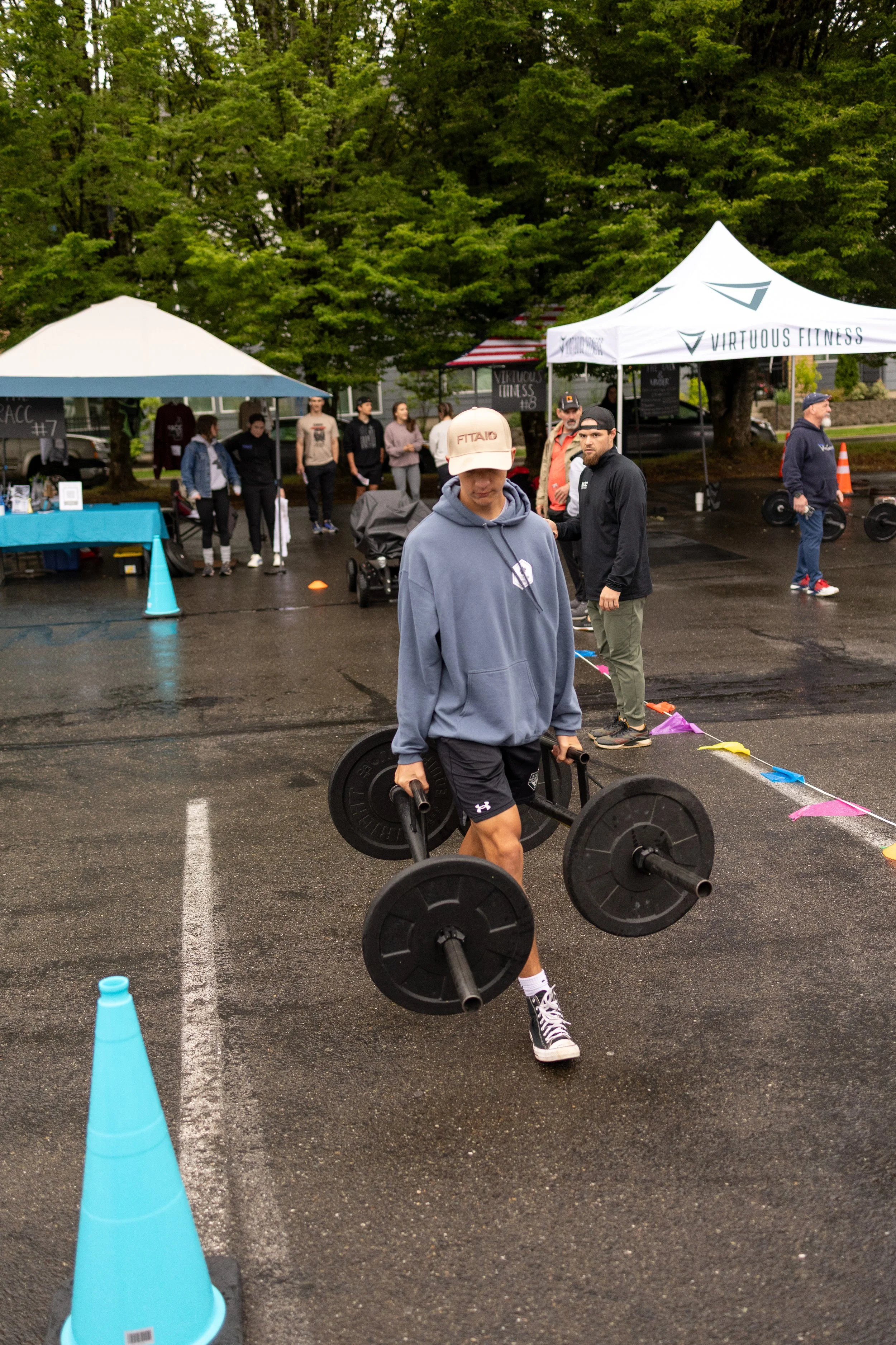 A young man in a hoodie and shorts walking on wet pavement while carrying a barbell with weight plates on each end during an outdoor fitness event. Several people and tents are in the background.