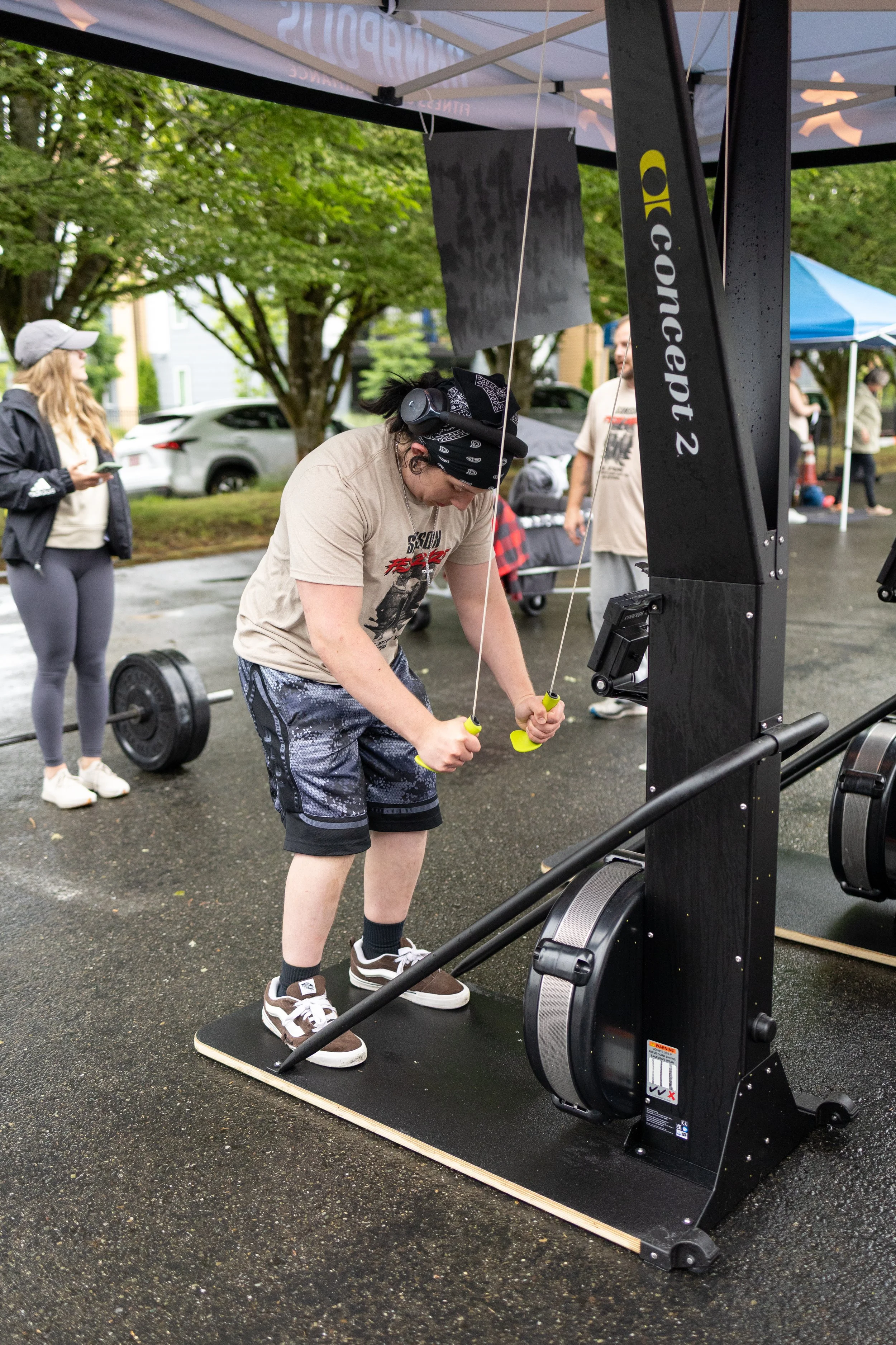 A person works out on a Concep1 2 elliptical trainer outdoors, with other people and tents in the background on a rainy day.