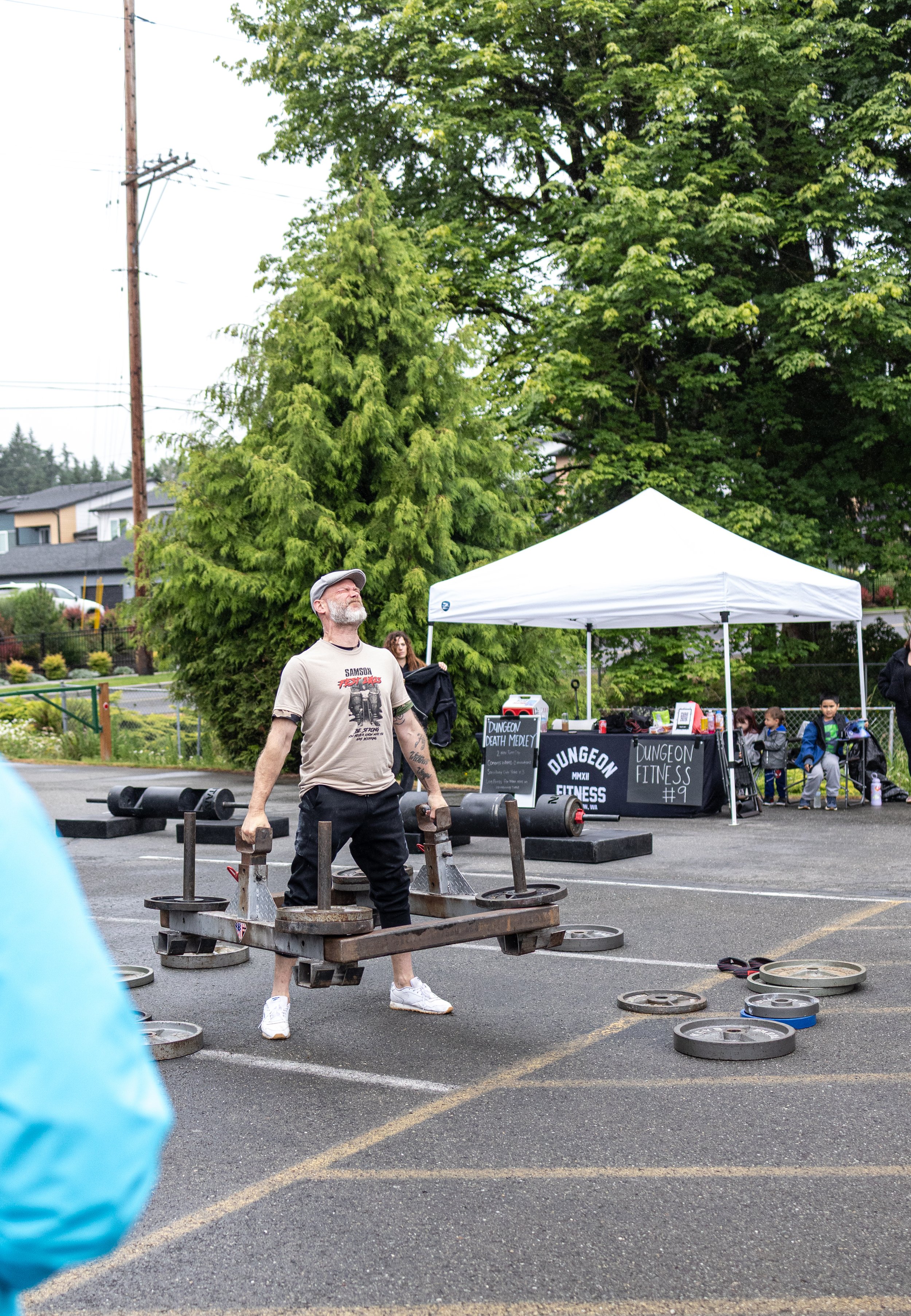 Man lifting heavy weight plates during an outdoor fitness event, with a small group of children and a table with fitness signs in the background.