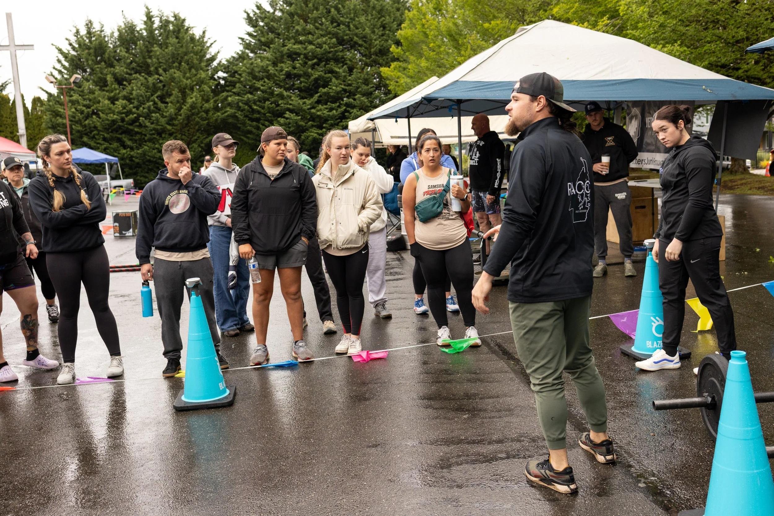 Group of people gathered outdoors on a rainy day, listening to a man in a black cap and jacket giving instructions, with tents and trees in the background.