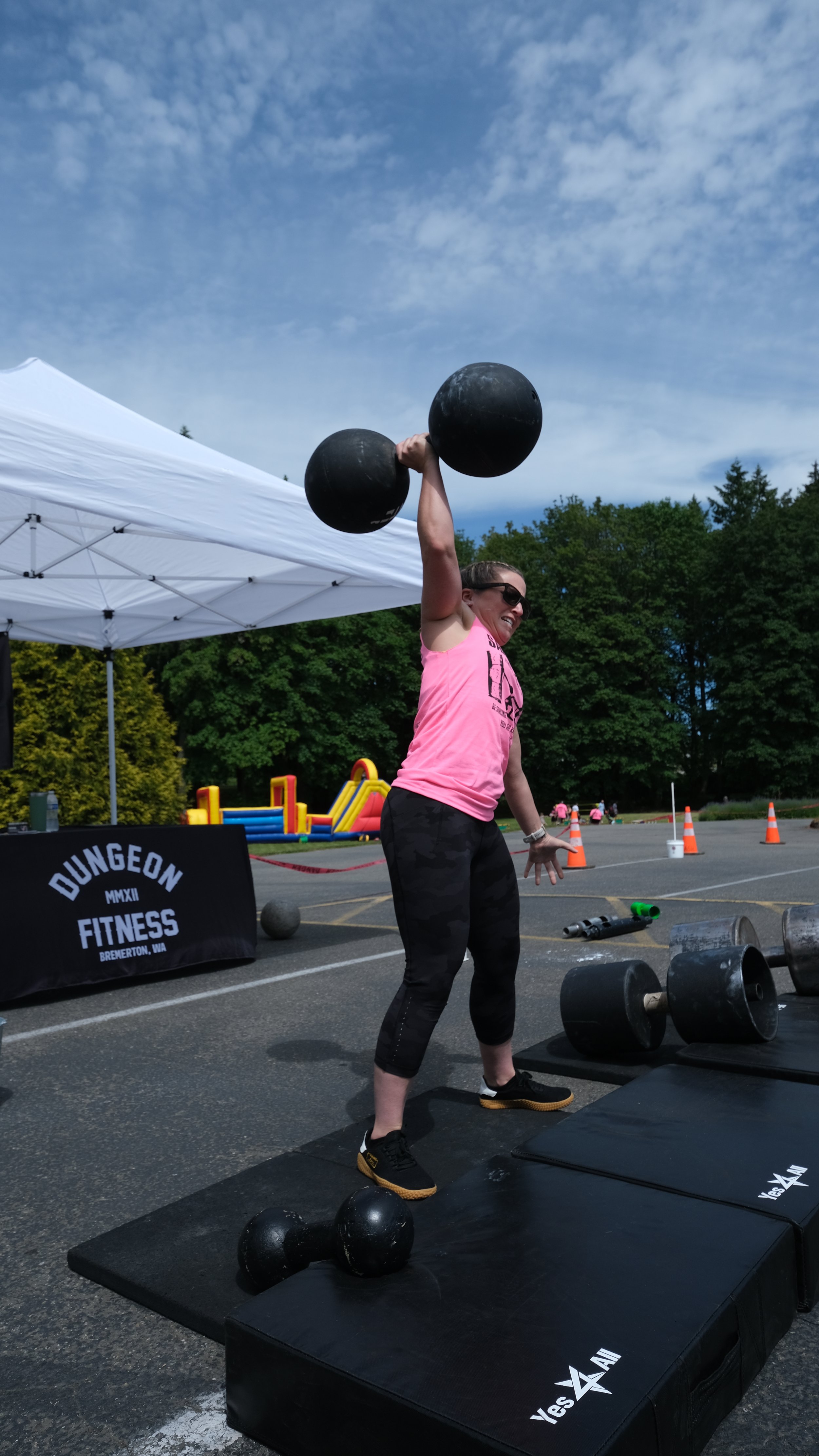 A person in a pink tank top lifting a large dumbbell overhead at an outdoor fitness event, with a white tent and trees in the background.