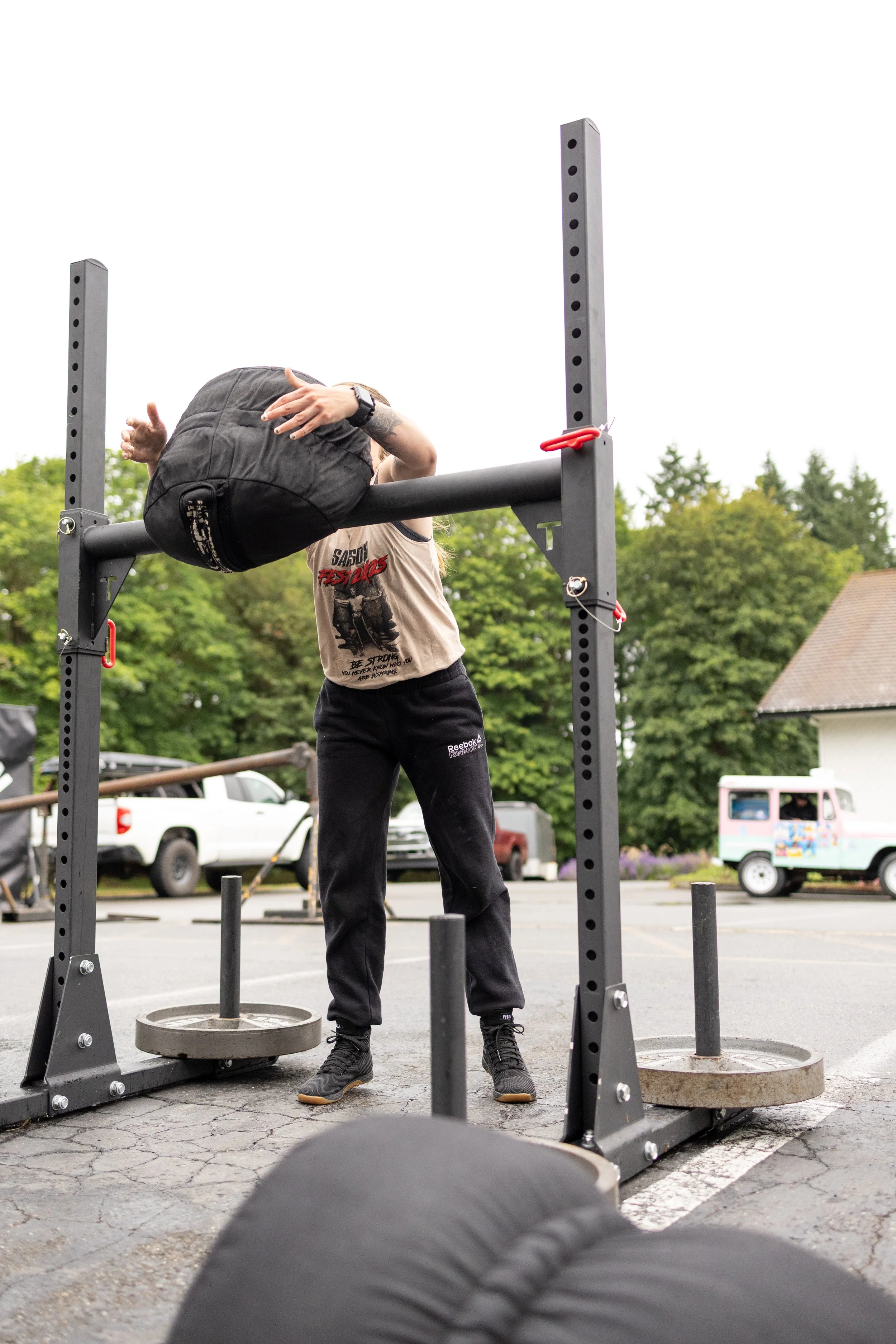 A person lifting a heavy sandbag onto a weightlifting platform outdoors, with a parking lot and trees in the background.