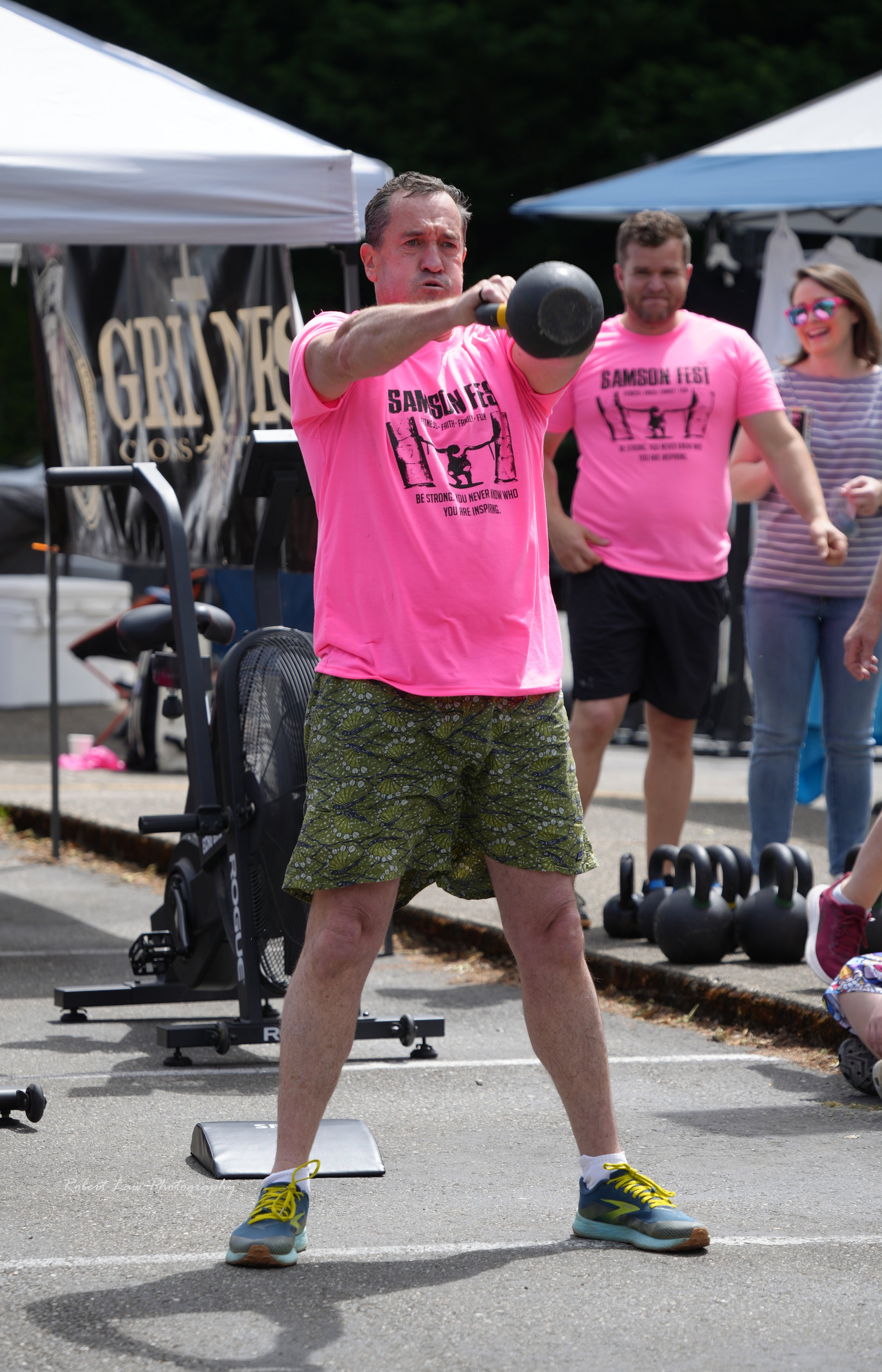 Man exercising with kettlebell at outdoor fitness event, wearing pink Samson Fest shirt and patterned shorts.