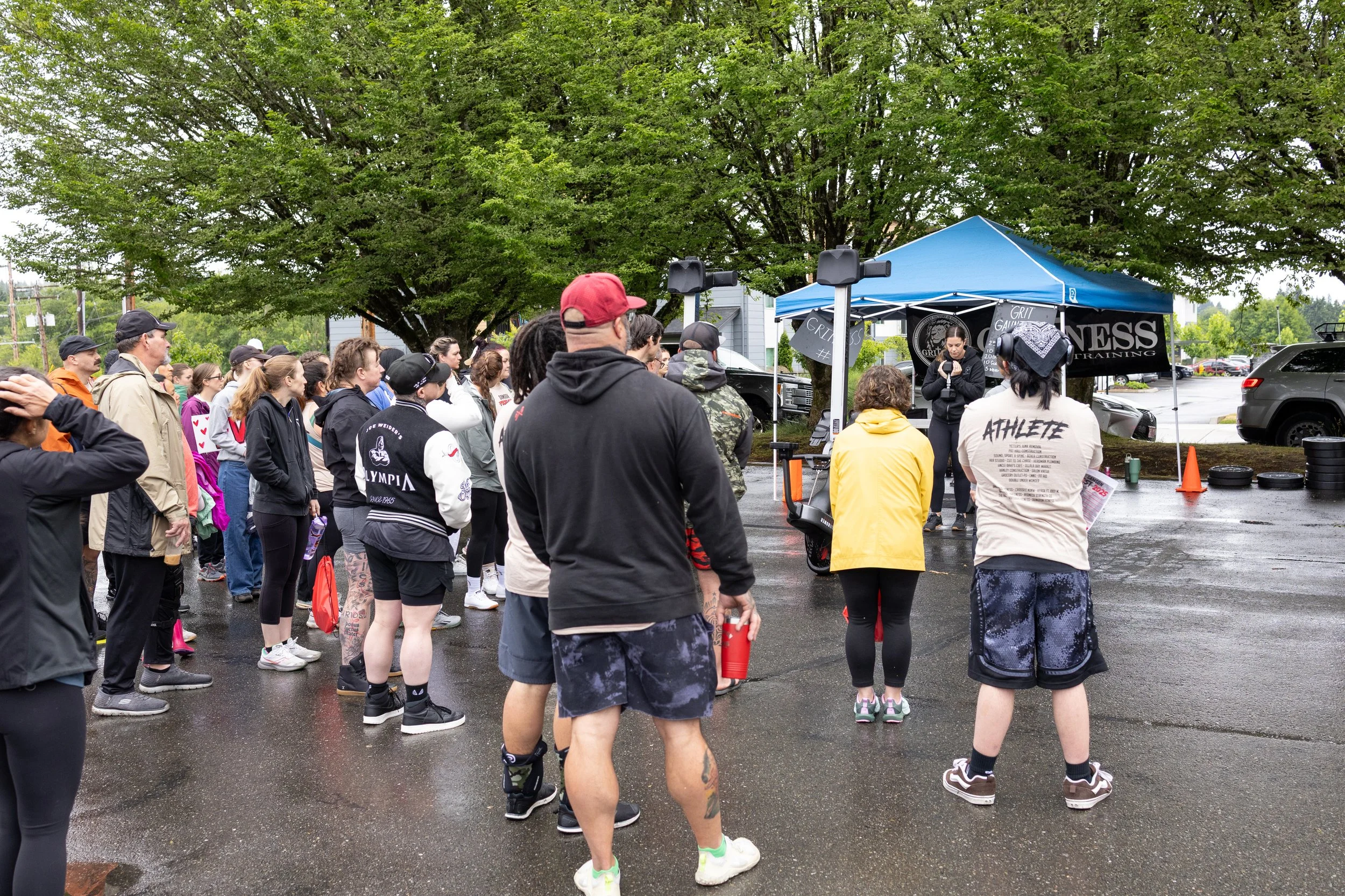 Group of people gathered outdoors on wet pavement, listening to a speaker under a blue canopy tent with fitness branding, large trees in the background.