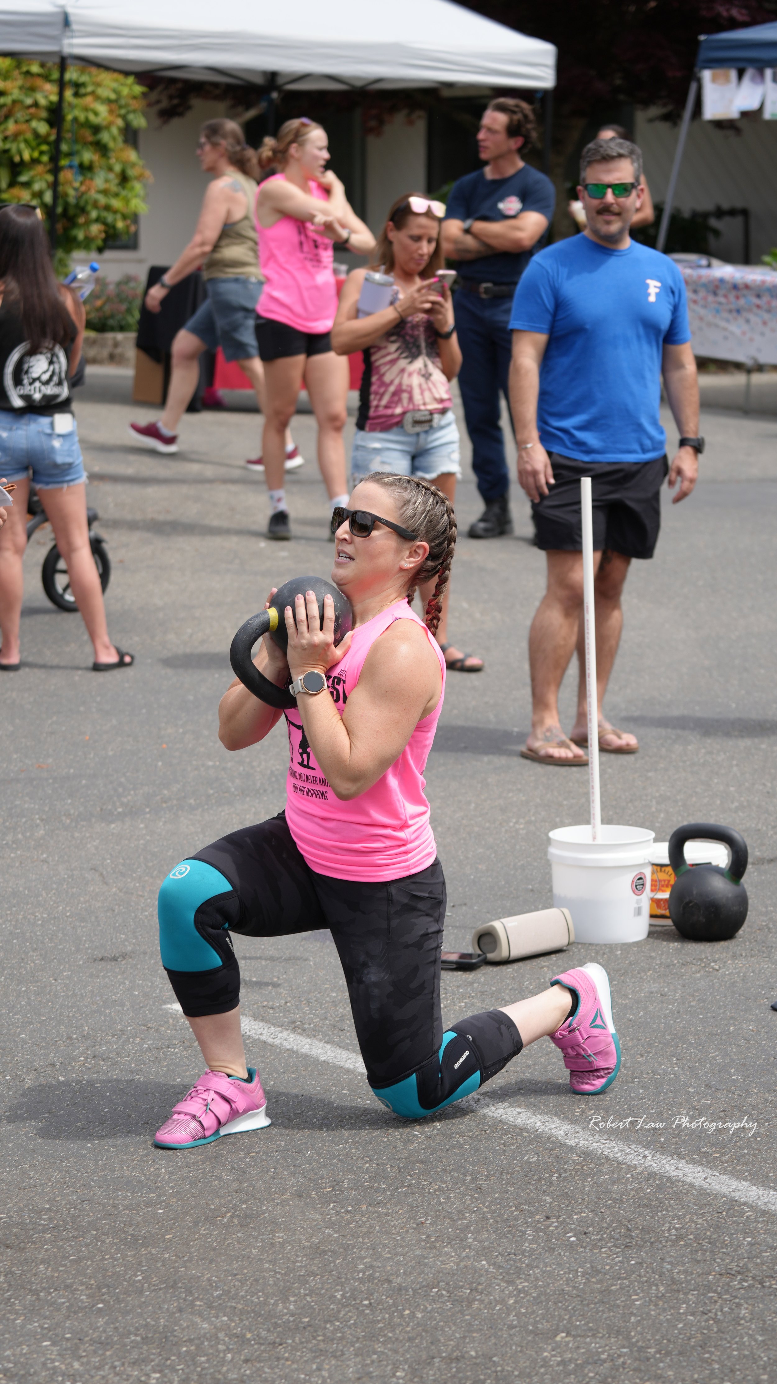 Woman performing a lunge exercise with a kettlebell at an outdoor fitness event, wearing a pink tank top and black leggings, surrounded by people, and fitness equipment visible on asphalt.