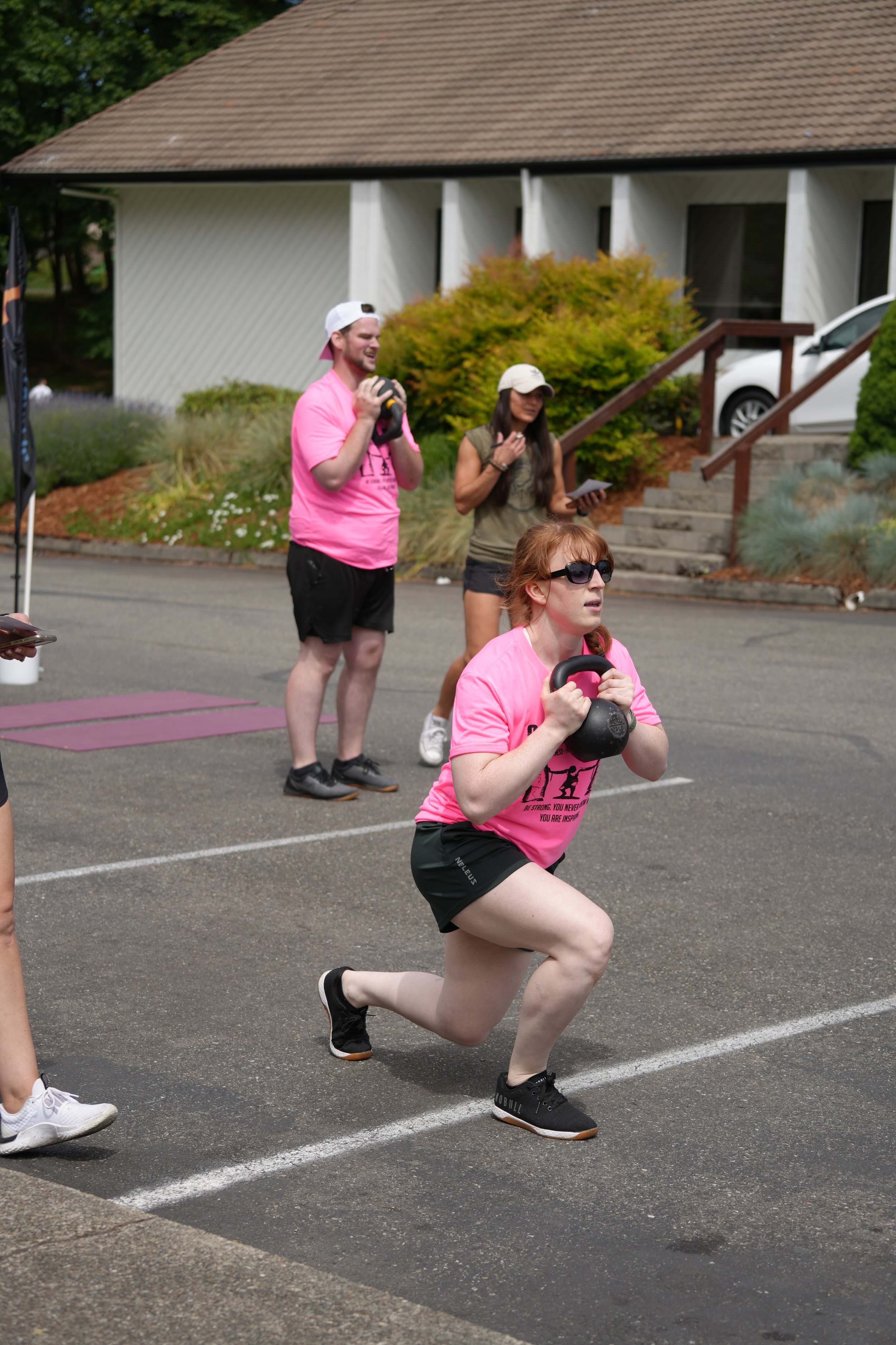 People in outdoor exercise class holding kettlebells, wearing pink shirts, concrete driveway, building and stairs in background.