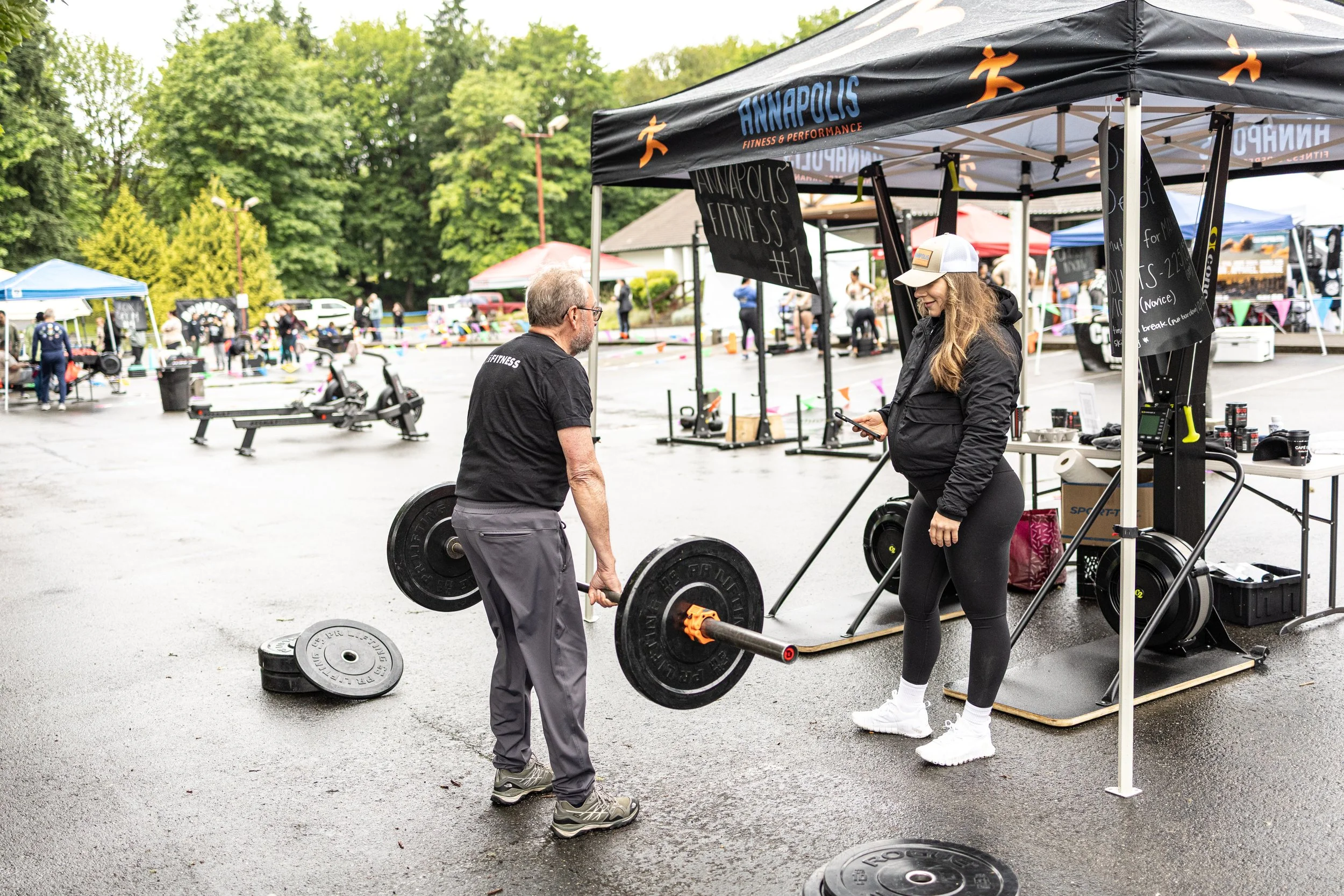 A man lifting a barbell at an outdoor fitness event, with a woman in athletic clothing standing nearby under a tent that says 'Annapolis Fitness & Performance'.