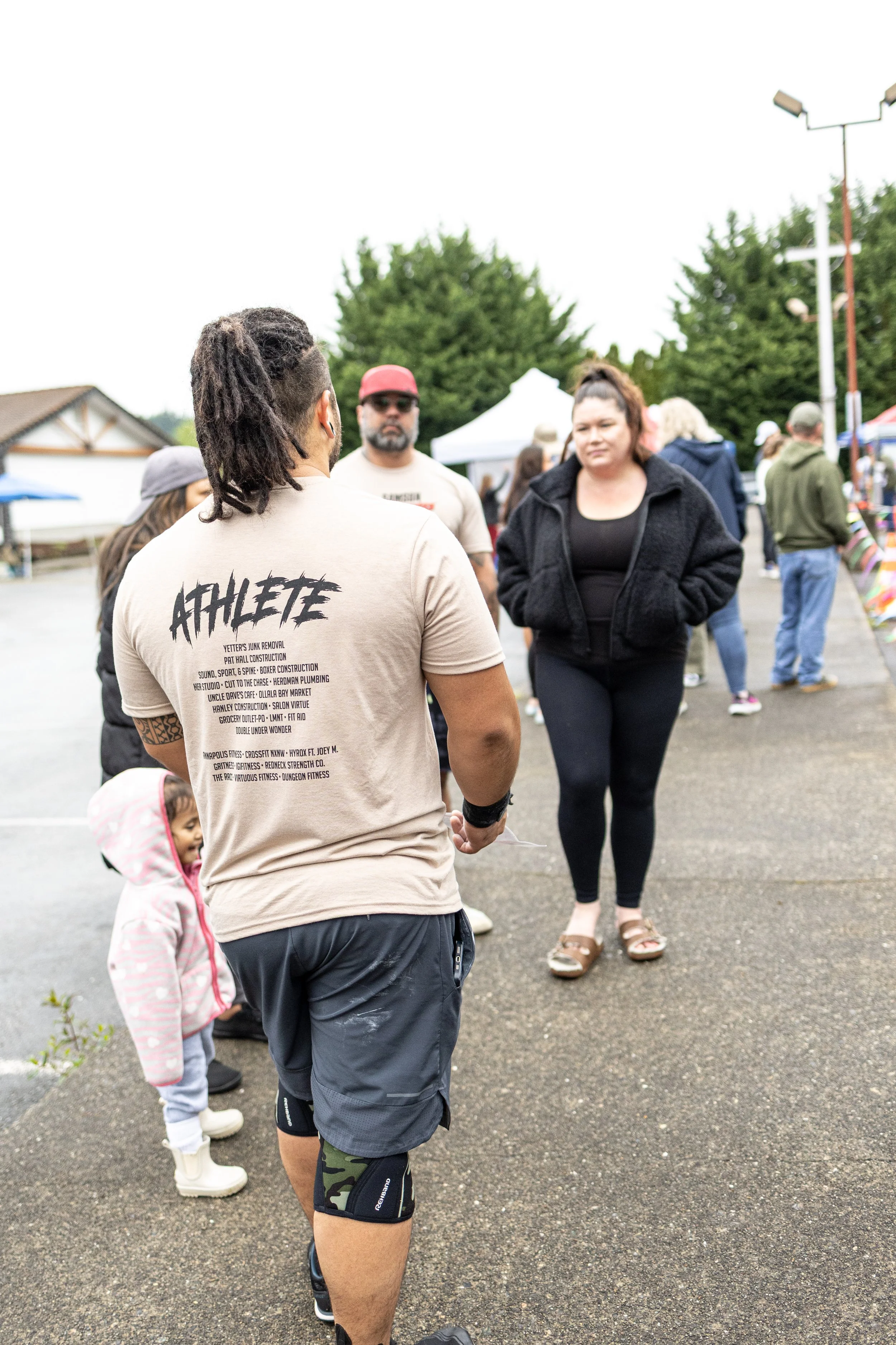 People walking outdoors at a market or fair on a cloudy day, with tents and stalls in the background.