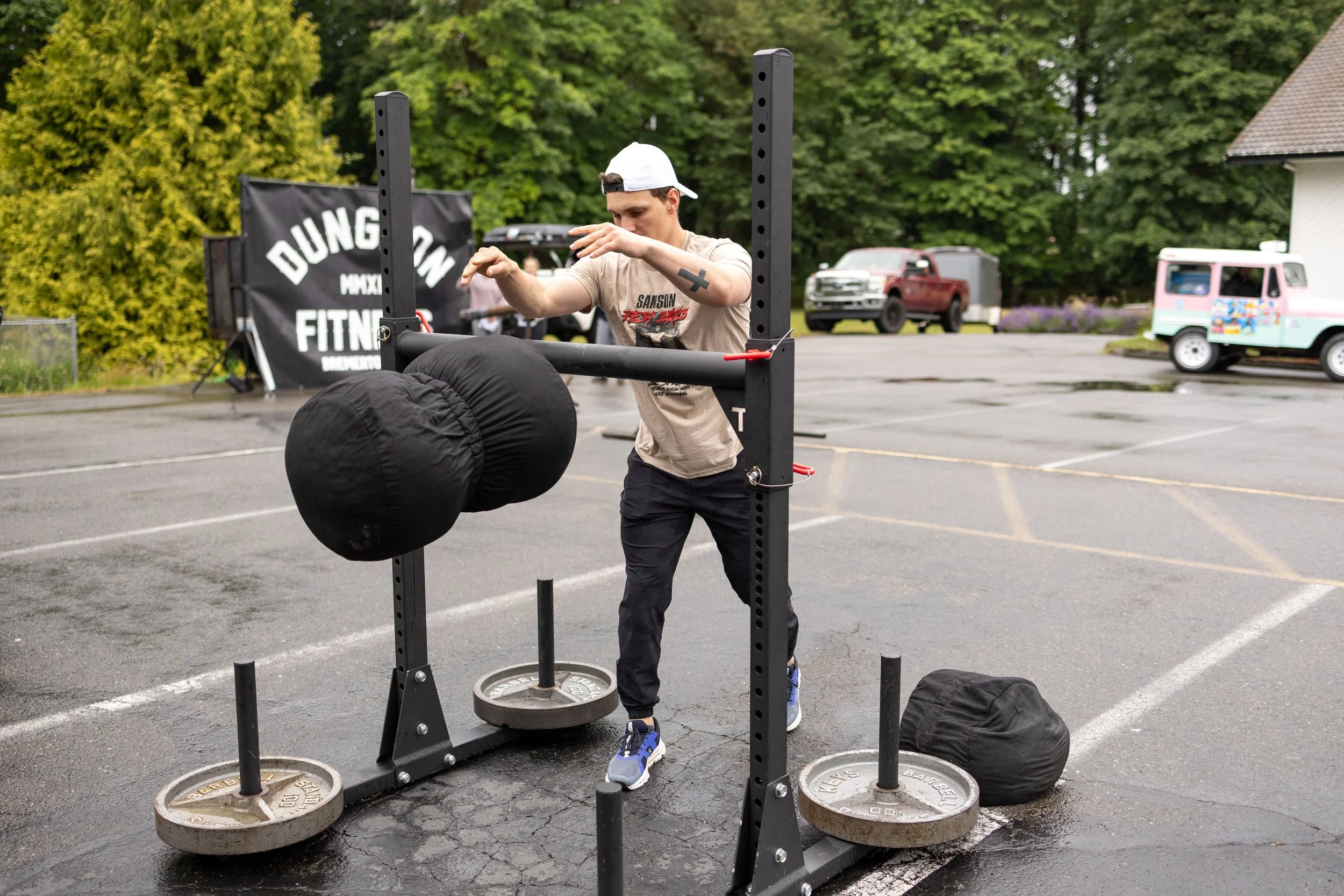 A person wearing a beige T-shirt, black pants, blue sneakers, and a white cap is working out with a sled push or pull station in an outdoor parking lot. There are large black cylindrical weights attached to the station, and a black bag on the ground 