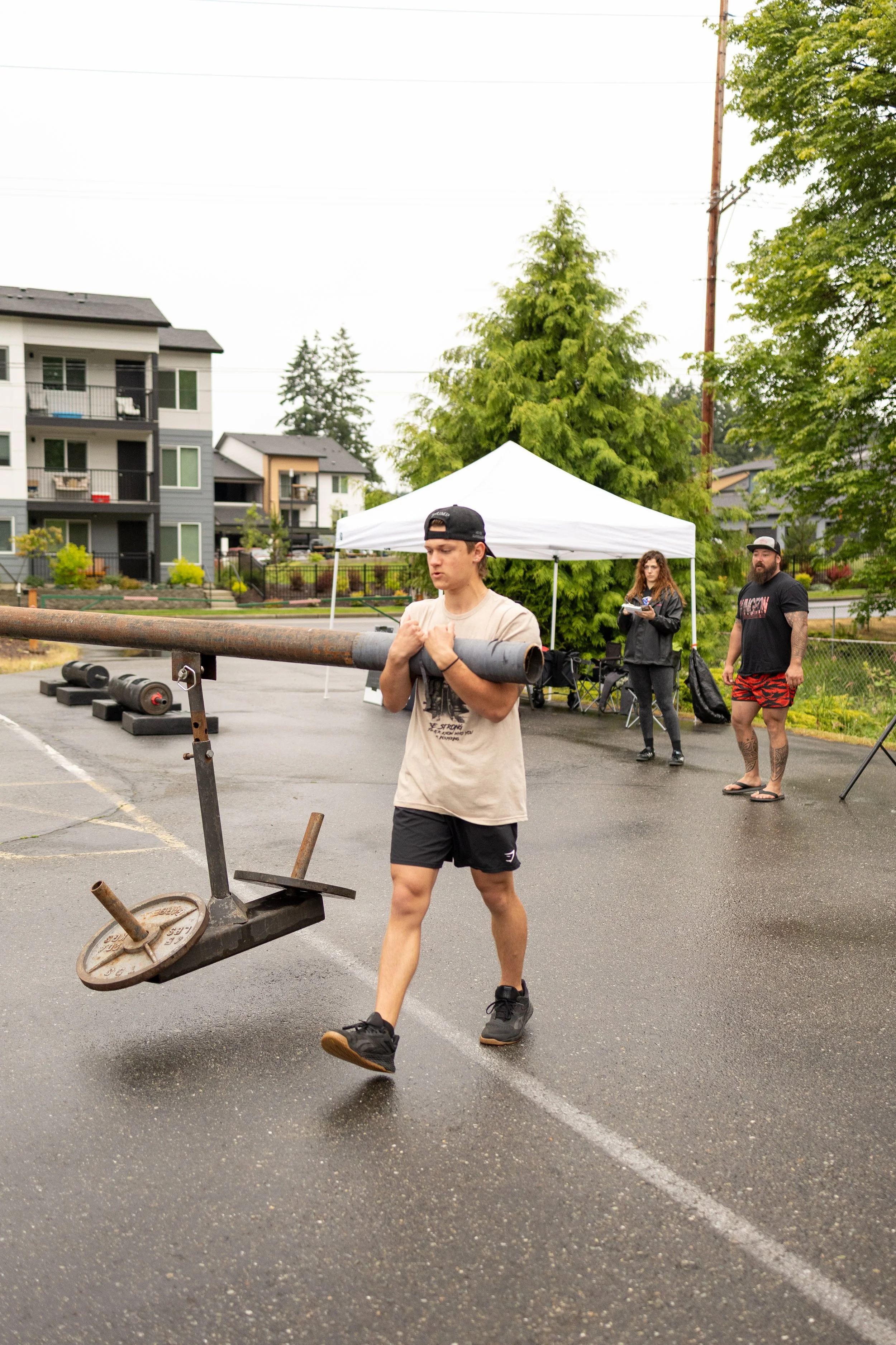 Young man in beige t-shirt and black shorts carrying a large metal bar on his shoulders during an outdoor weightlifting event, with a woman and man in casual clothes standing under a white canopy in the background on a rainy day in a parking lot.