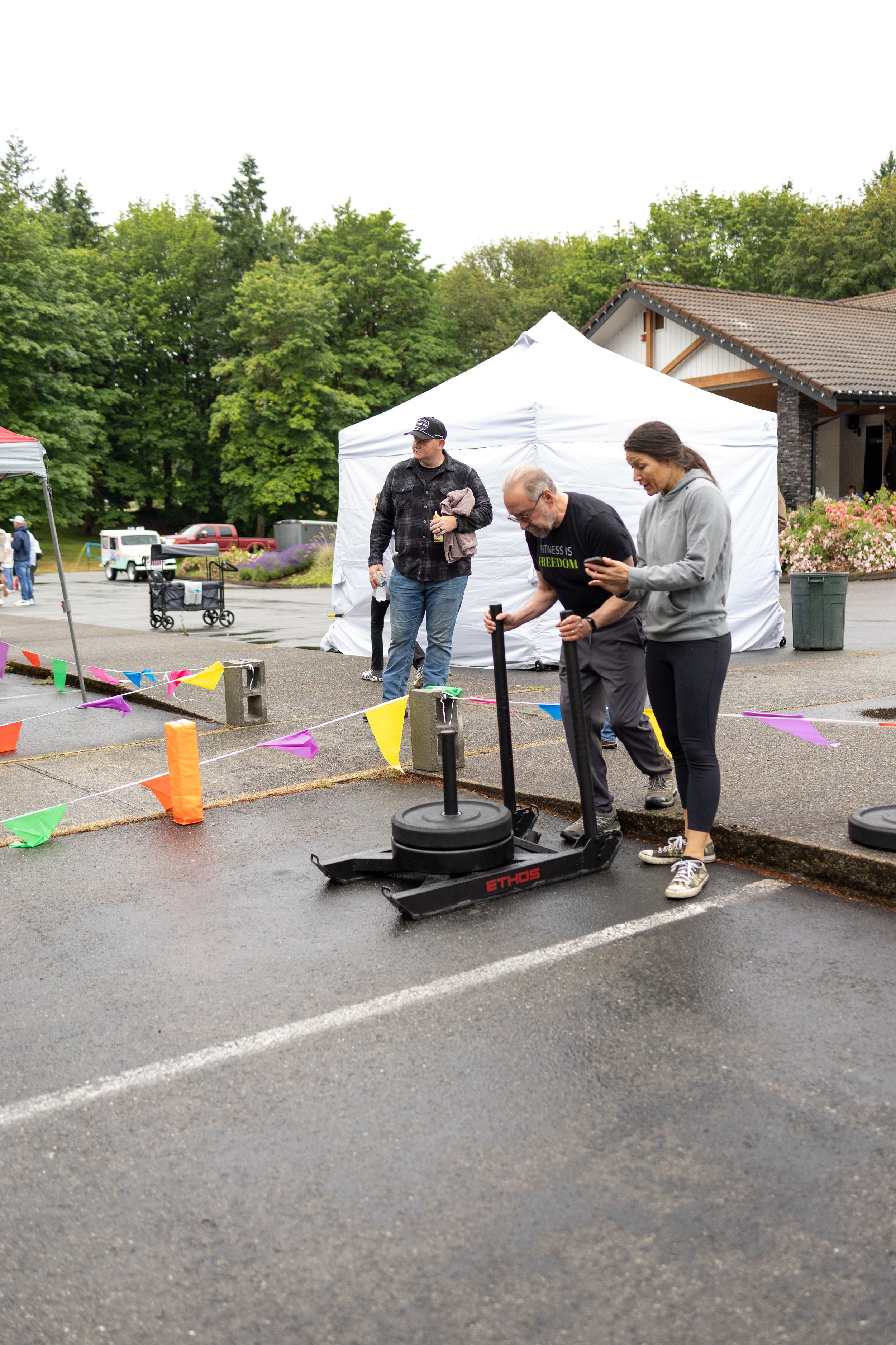 People preparing for a fitness event outdoors, with a man and woman placing weights on a sled, surrounded by colorful flags and tents, on a wet parking lot.