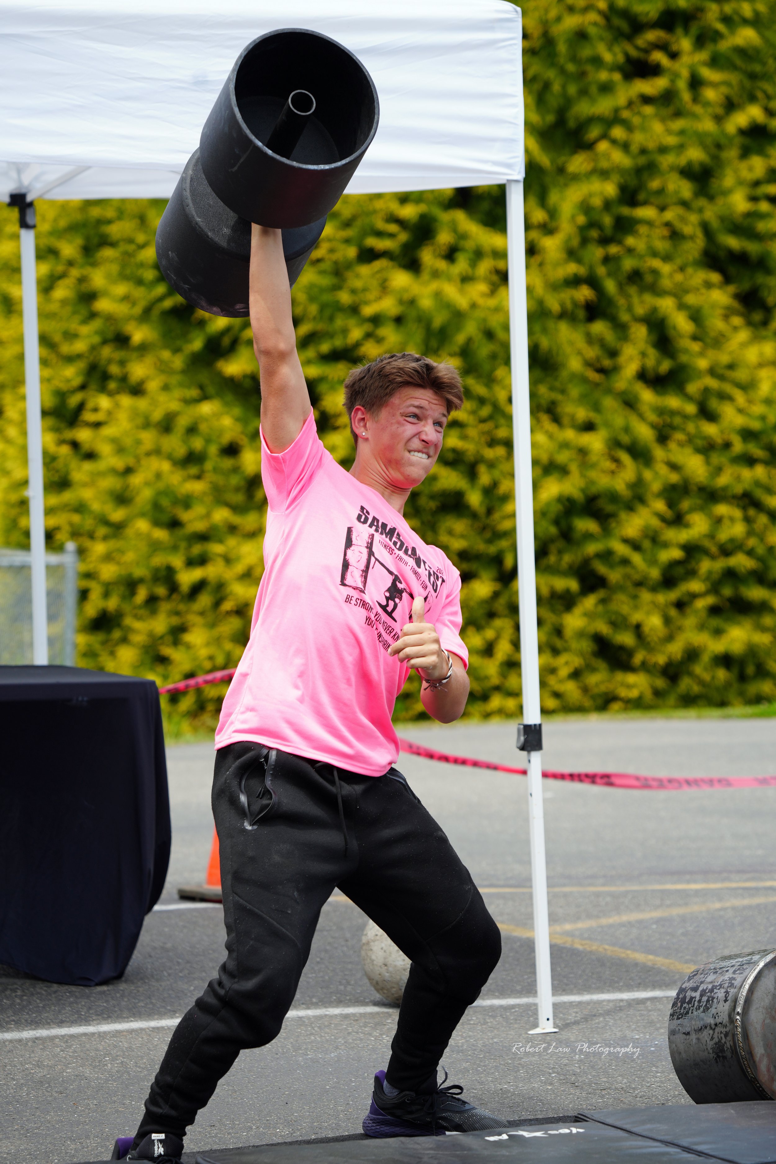 Person in a pink T-shirt and black pants lifting a heavy weight overhead during an outdoor strongman competition.