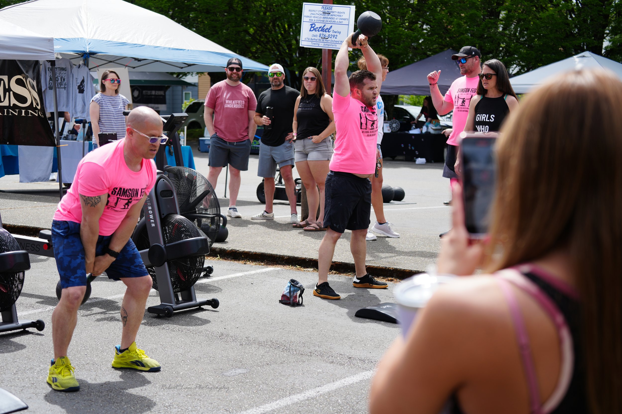 Outdoor fitness event with participants exercising, lifting weights, and spectators watching in a parking lot.