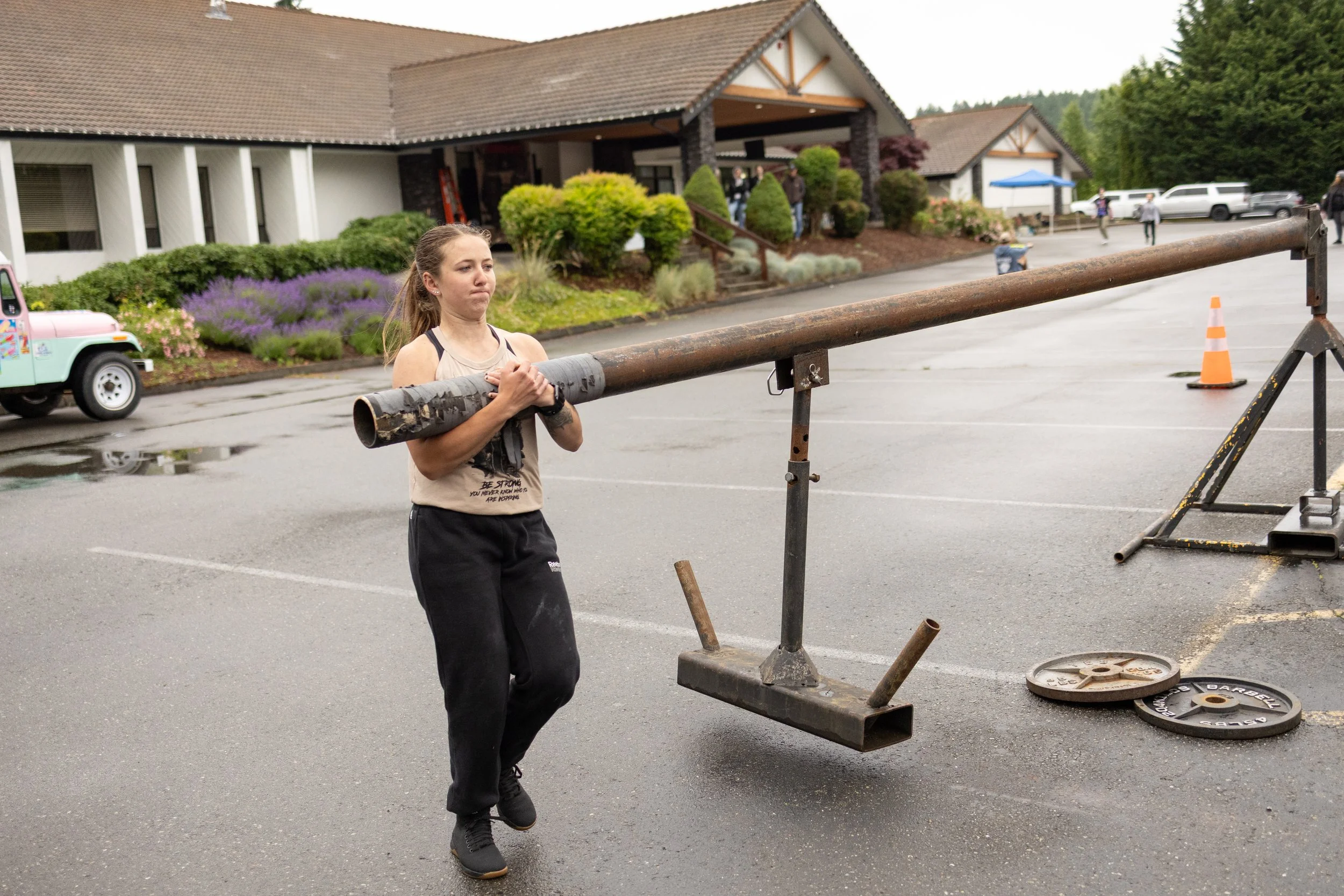 A young woman is lifting a large metal pipe in a parking lot, with weight plates and a cone nearby, in front of a building with shrubs and trees.