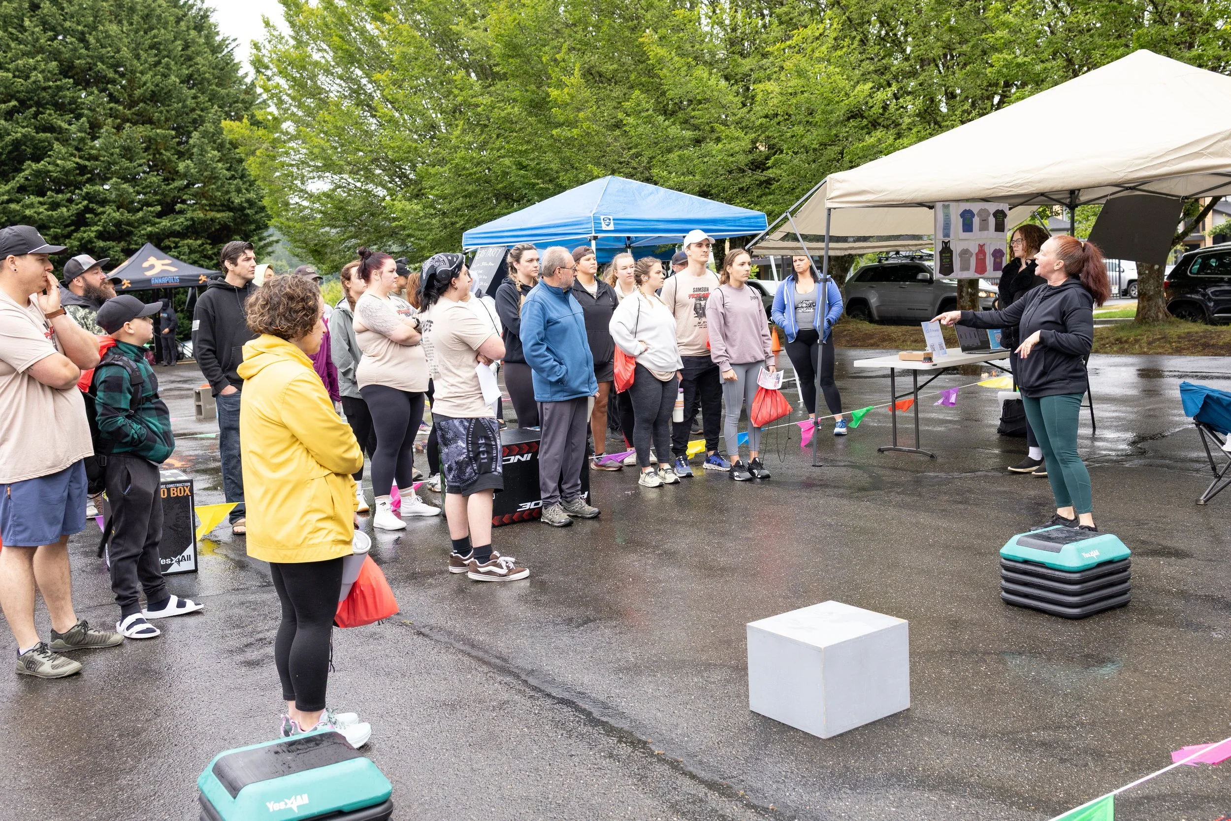 People gathered outdoors on a rainy day listening to a woman speak at an event with tents and banners.