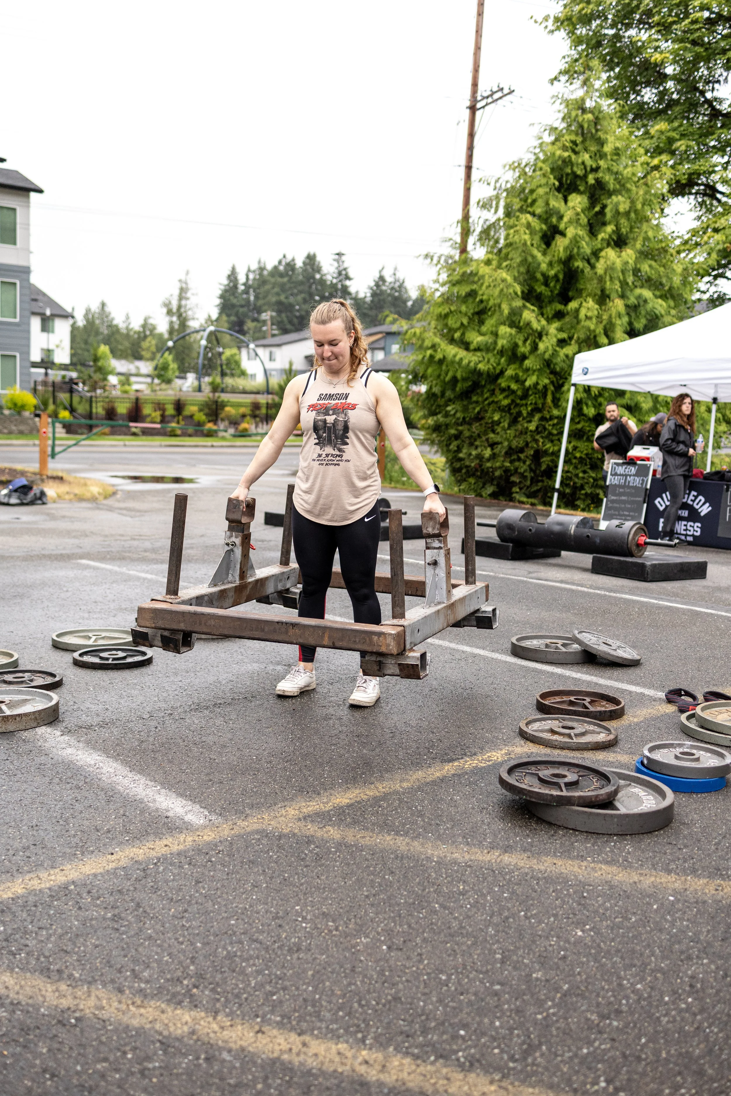 A woman lifting a wooden frame with weights on a parking lot during a fitness event, with other participants and equipment visible in the background.