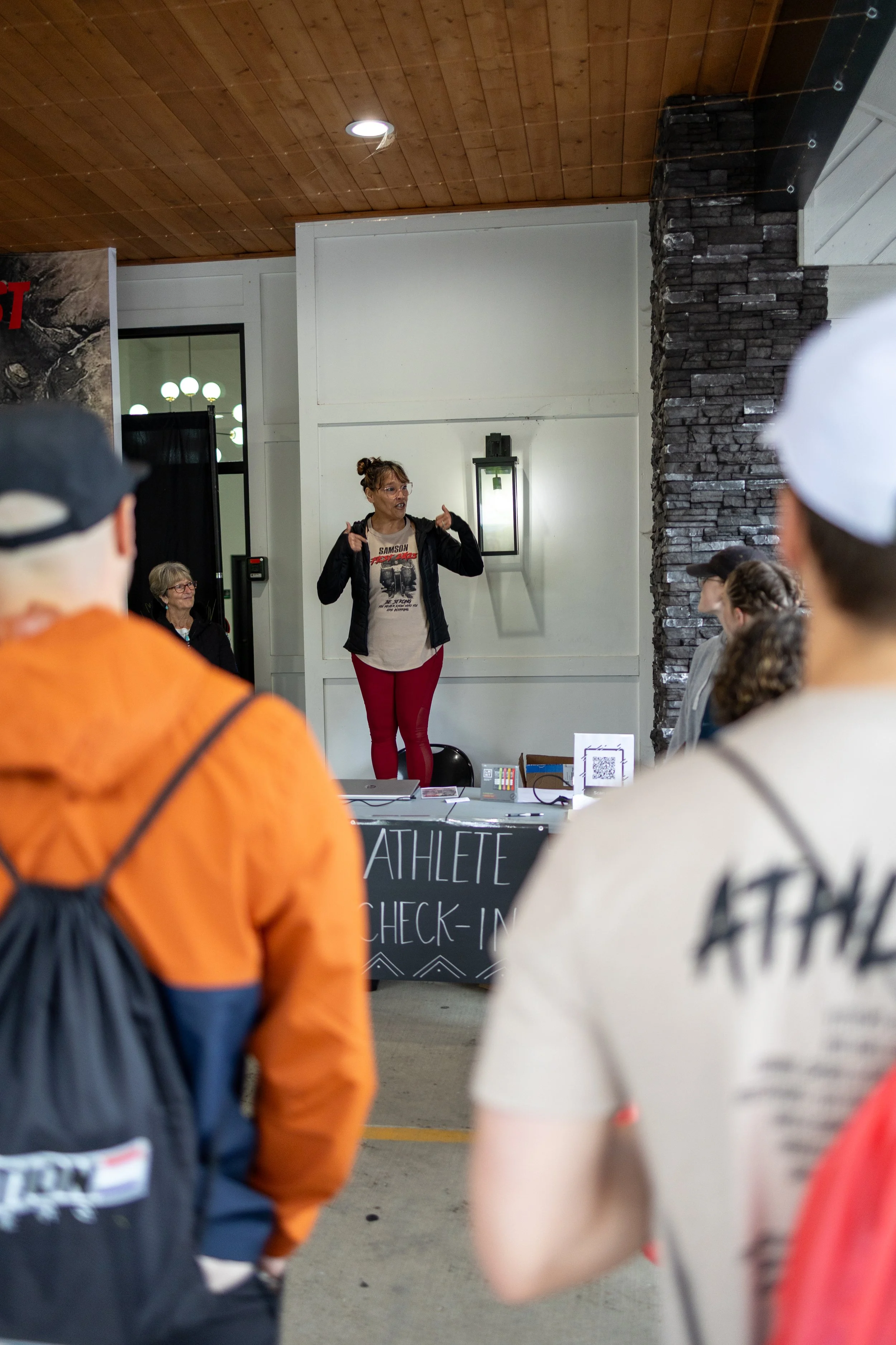 A woman stand at a check-in desk labeled 'ATHLETE CHECK-IN' speaking to a group of people inside a room with a wooden ceiling and stone wall.