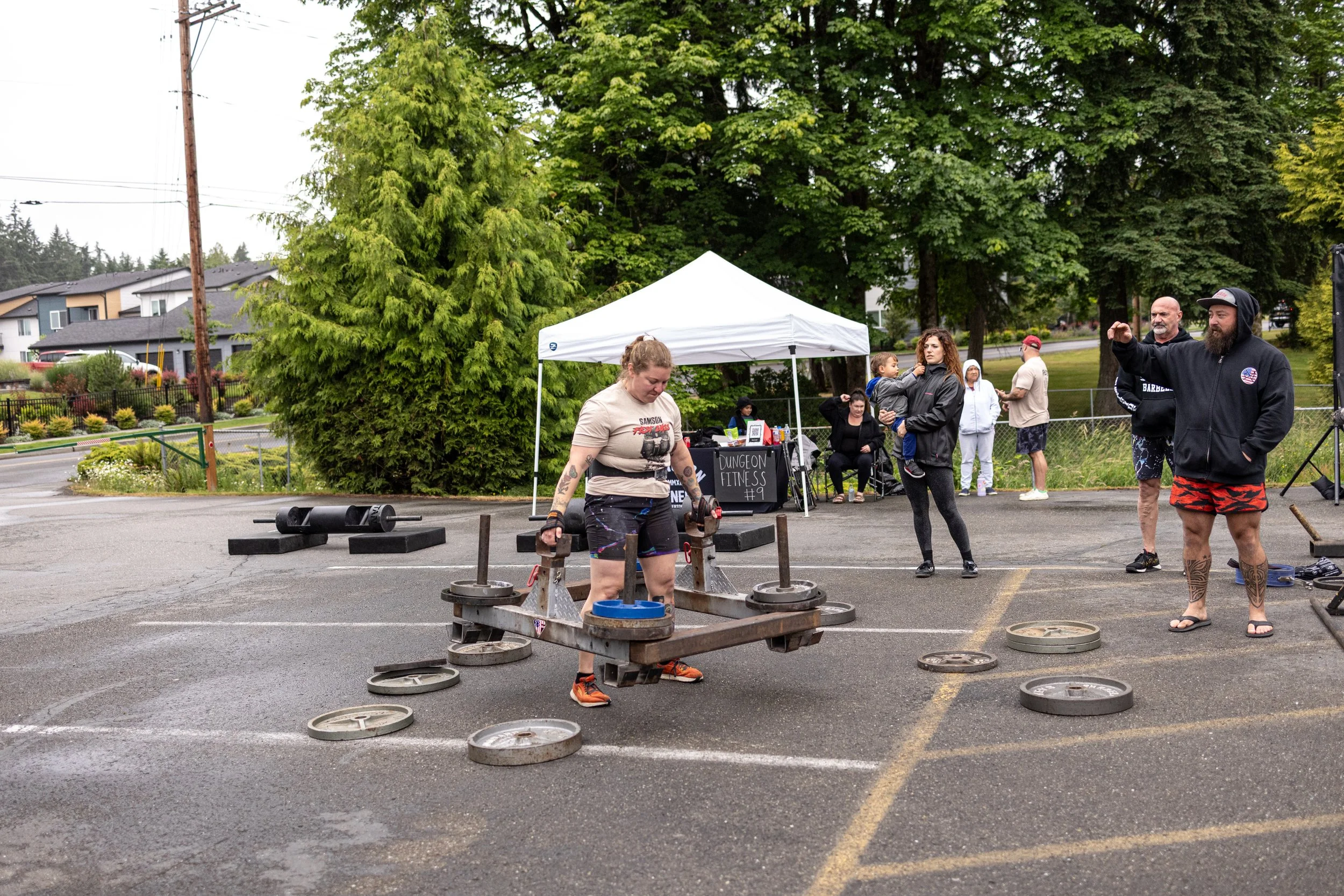 Woman lifting a barbell with weights at an outdoor fitness event, with spectators and a tent in the background.