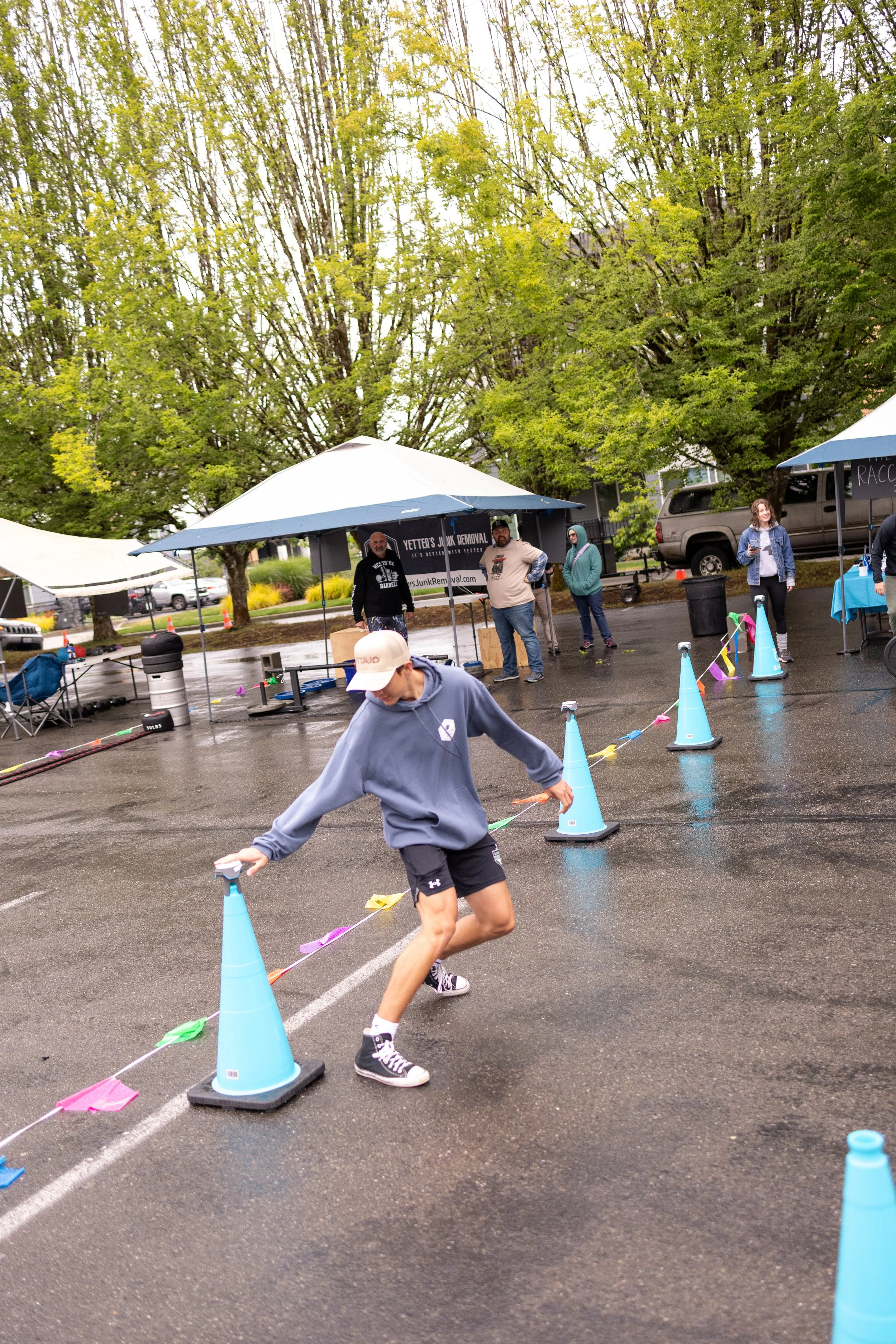 A young man running around orange and blue cones on a wet parking lot during a rainy day, with tents and people in the background.