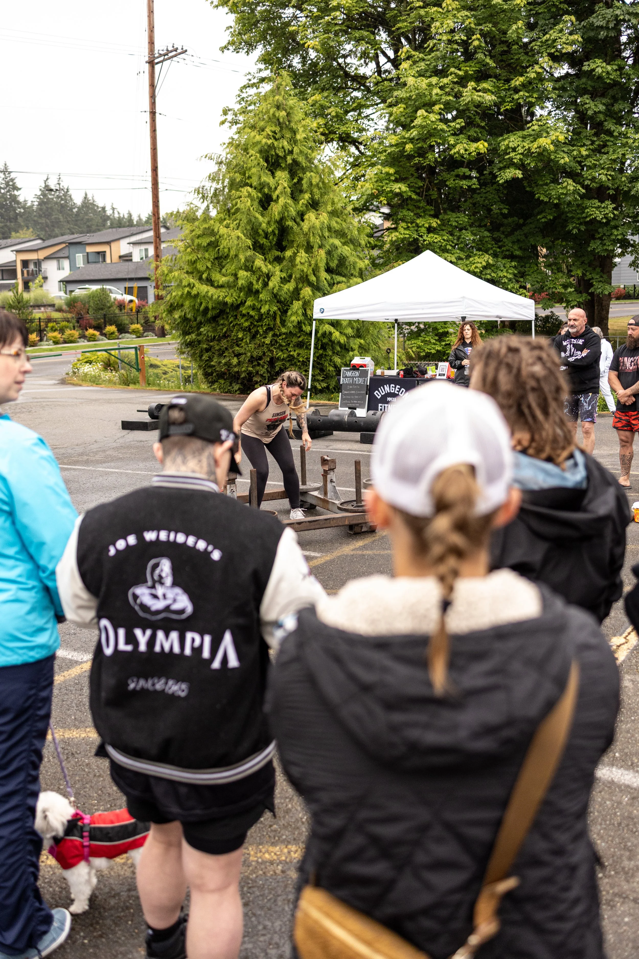 A woman participating in a strongman event lifts a heavy wooden log while several onlookers and a dog watch in a parking lot with trees and houses in the background.