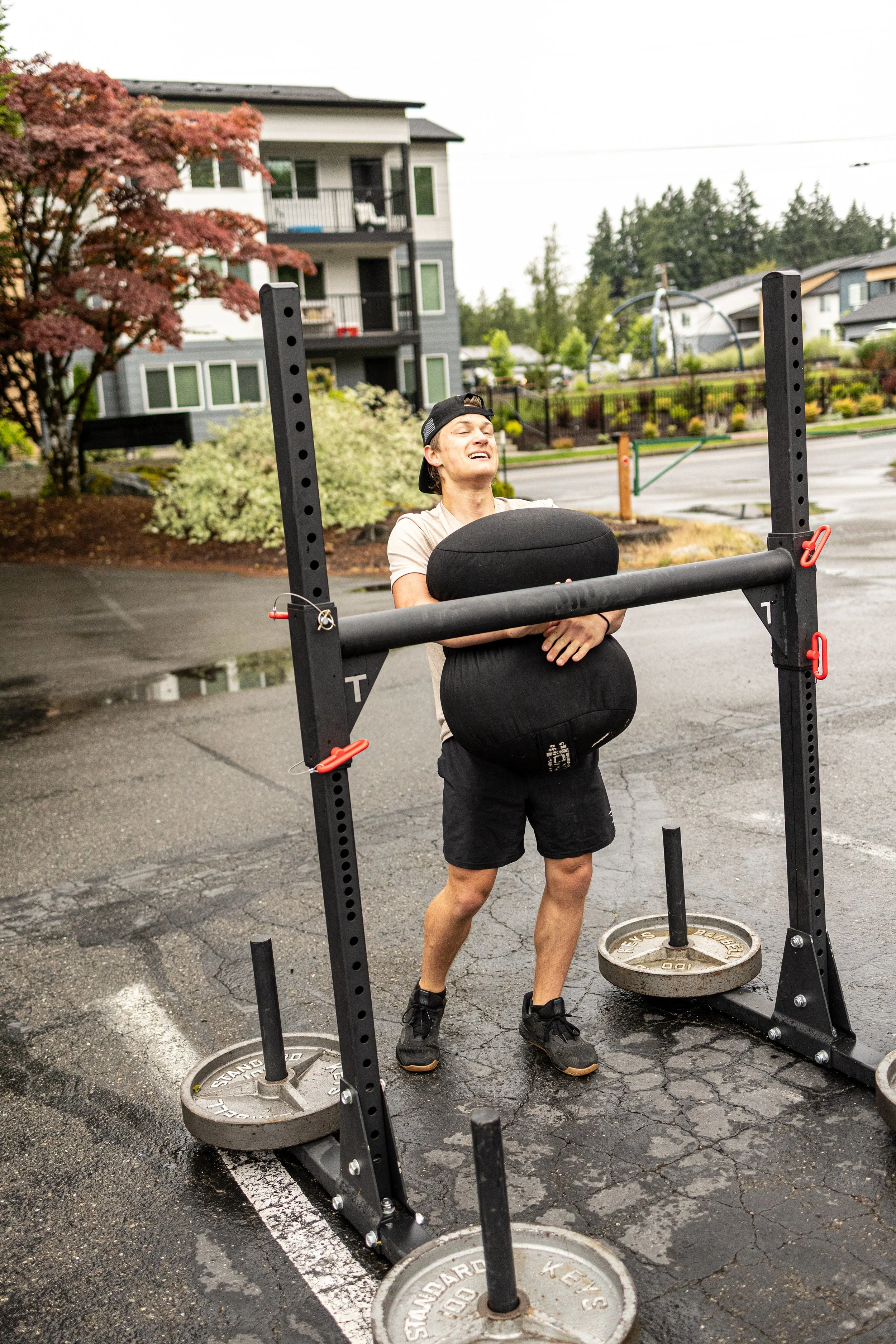 A person holding a large black weight plate in front of a squat rack outdoors during rainy weather.