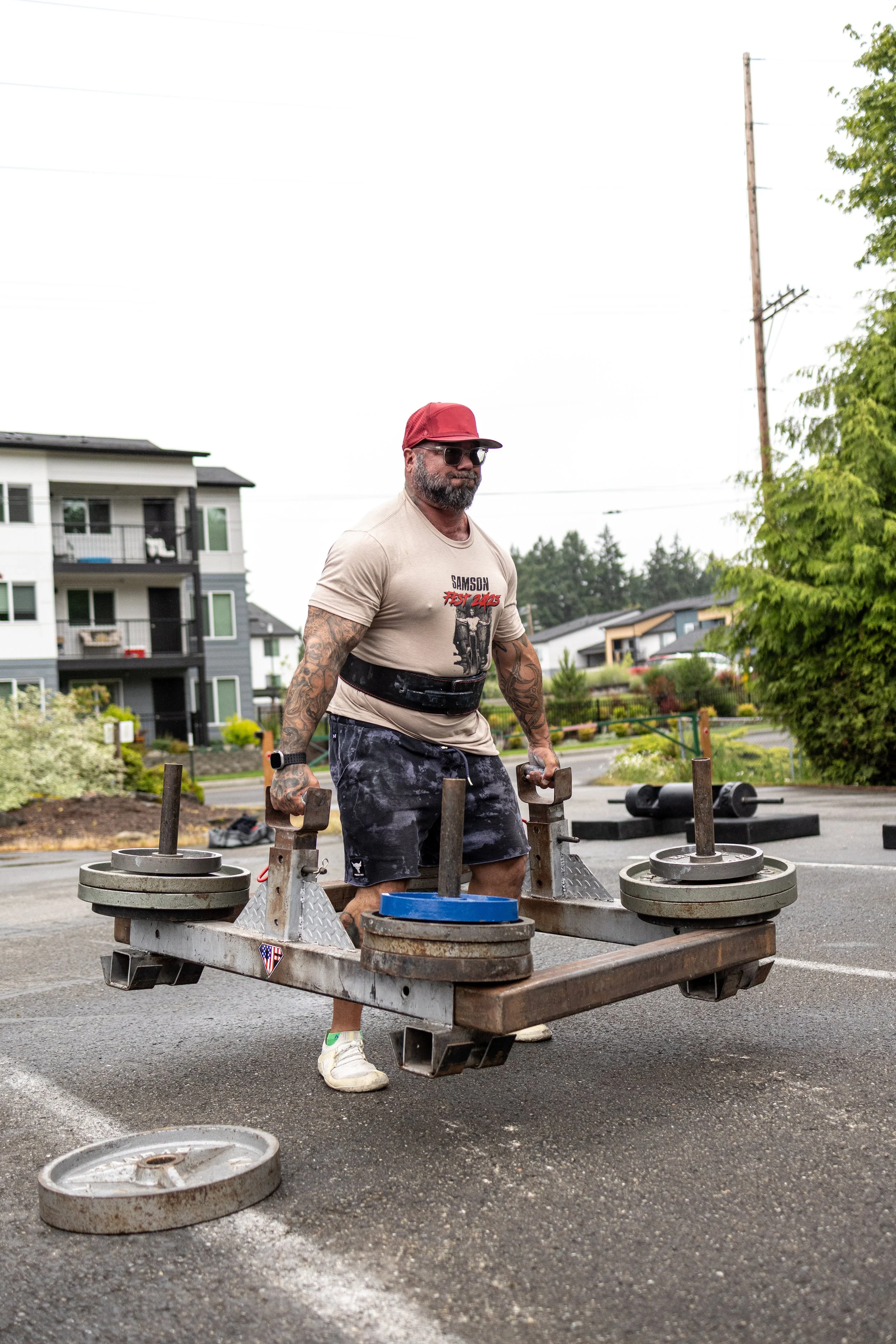 A man with tattoos, sunglasses, a red cap, Samson Fest T-shirt, lifting a weighted platform in an outdoor fitness competition in south kitsap for Samson Fest.