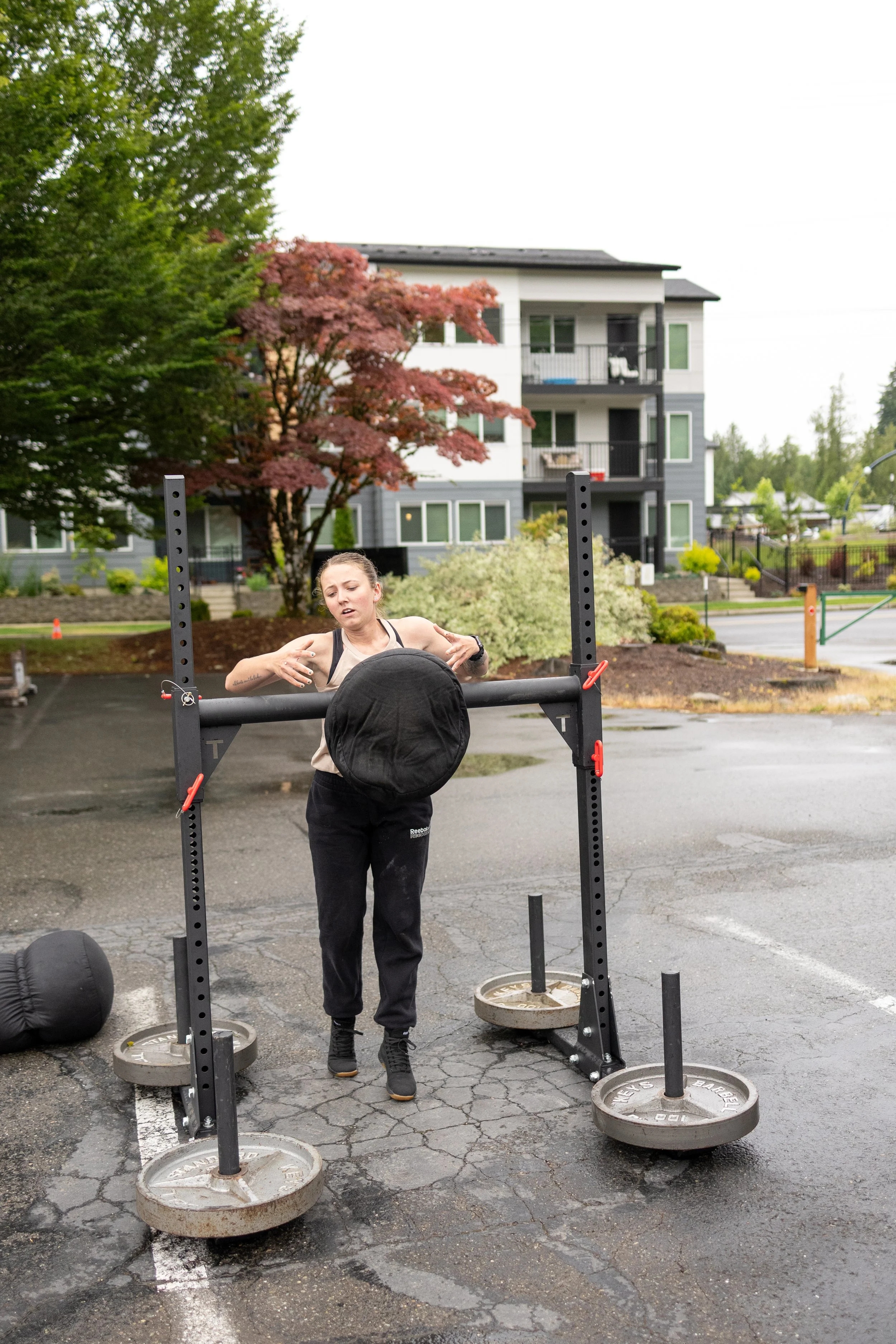 A woman exercising outdoors with a tire flip station in a parking lot, with residential buildings and trees in the background.