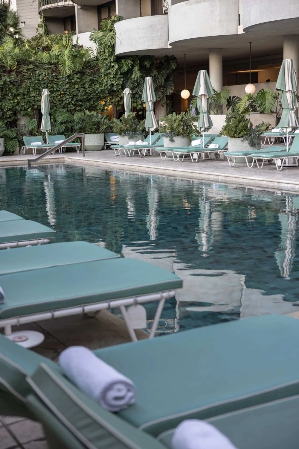 Empty poolside lounge chairs with rolled towels, umbrellas, and greenery surrounding a hotel swimming pool.