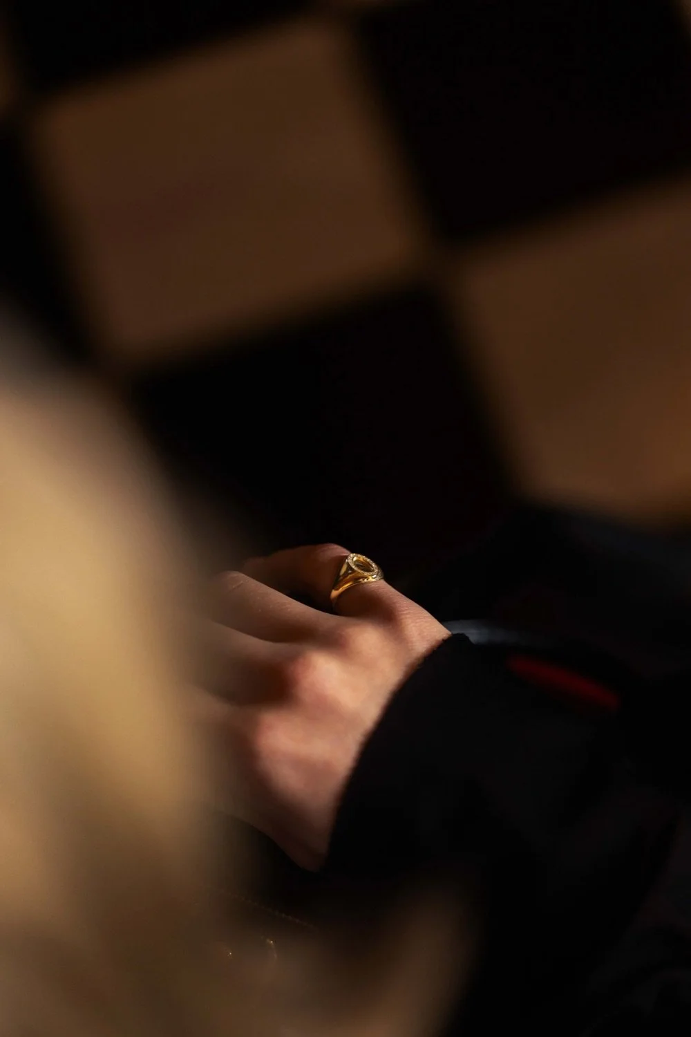 Close-up of a hand wearing a gold ring, resting on a surface with muted lighting.