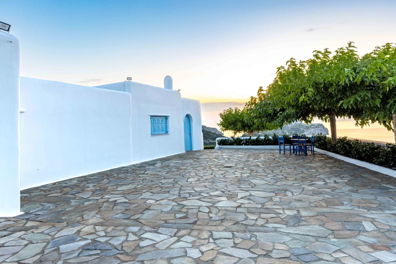 A white Mediterranean-style building with blue accents, a stone patio, large trees, and a view of the coastline at sunset.