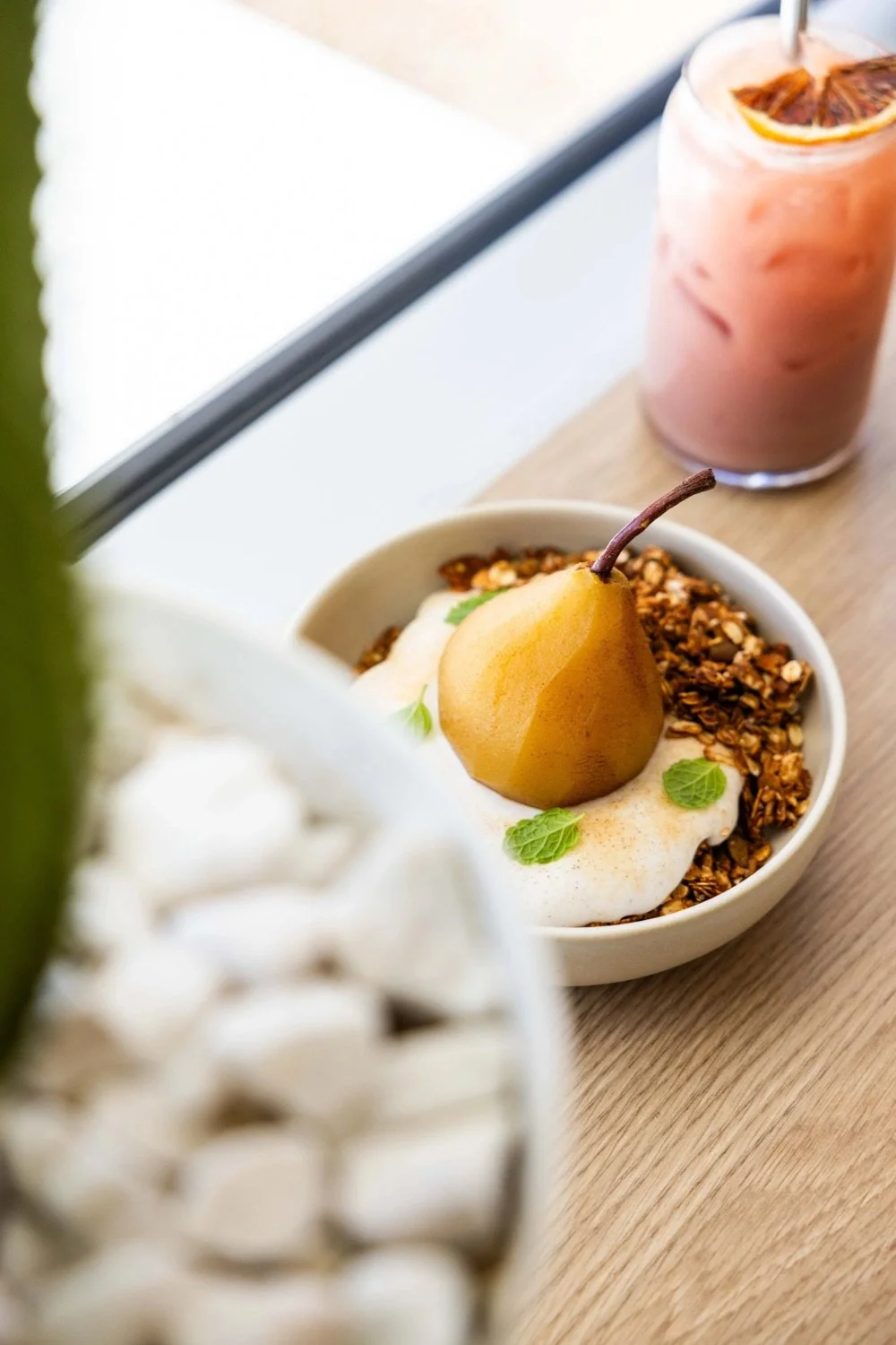 A bowl of yogurt topped with a poached pear, granola, and mint leaves, alongside a pink beverage with a lemon slice and a straw on a wooden table near a window.