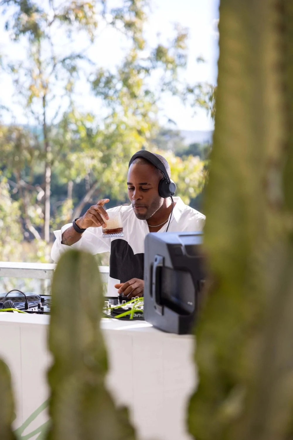 A man wearing a white shirt with black design, headphones, and a watch is standing behind a DJ mixing table outdoors, holding a glass with a beverage, with trees in the background.