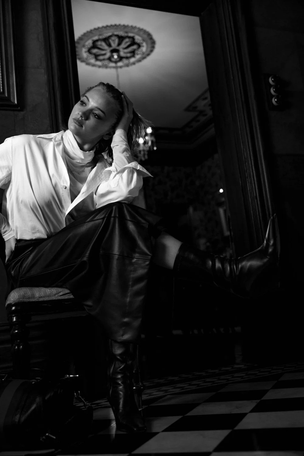 Black and white photo of a woman sitting on a chair indoors. She is wearing a white blouse, leather skirt, and knee-high boots. She has her head resting on her hand, and is looking to her left with a contemplative expression.